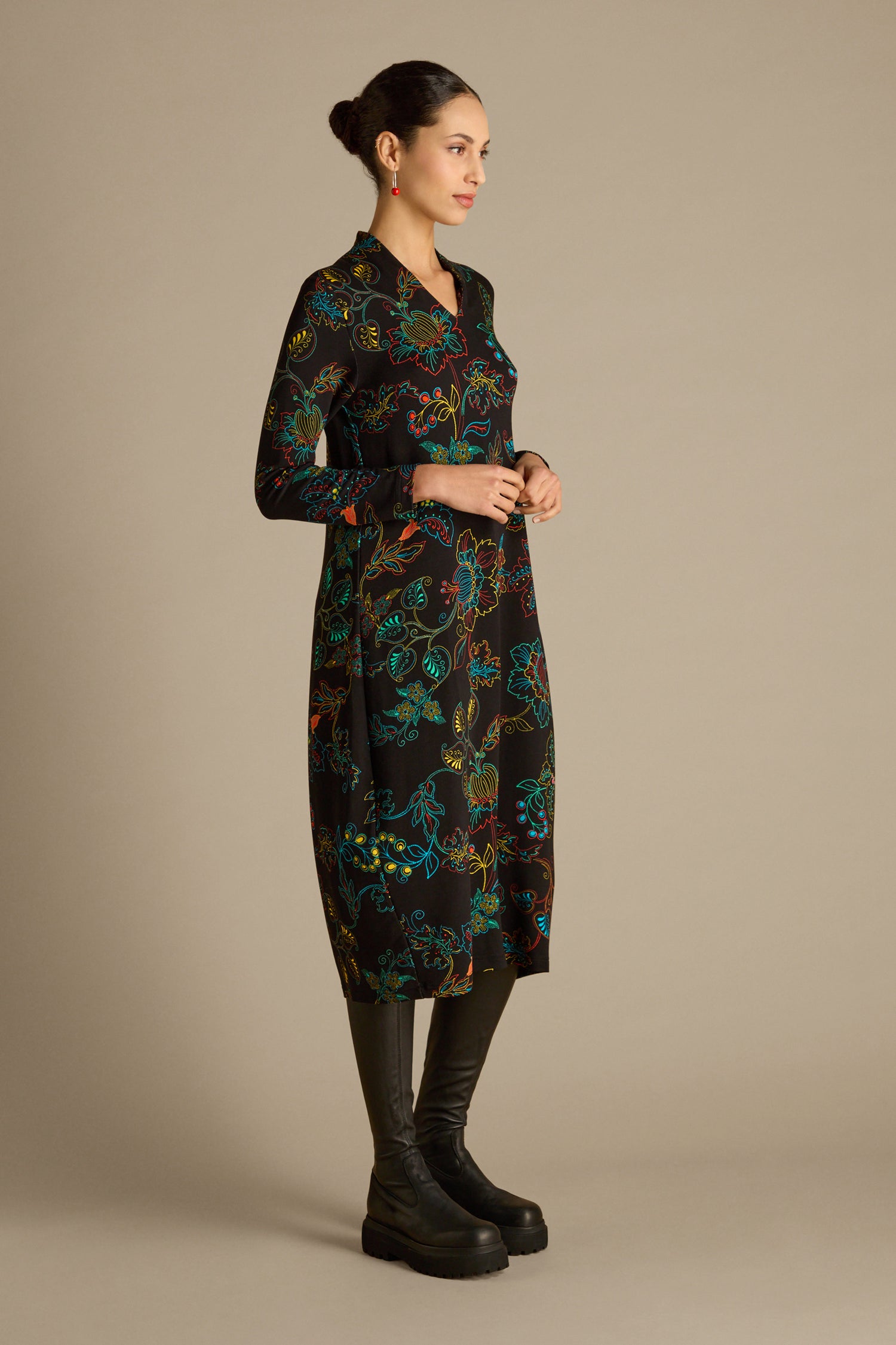 A woman wearing the Bright Dotted Floral Jersey Dress, featuring a V-neckline, pairs it with black boots as she stands against a neutral background, facing right.