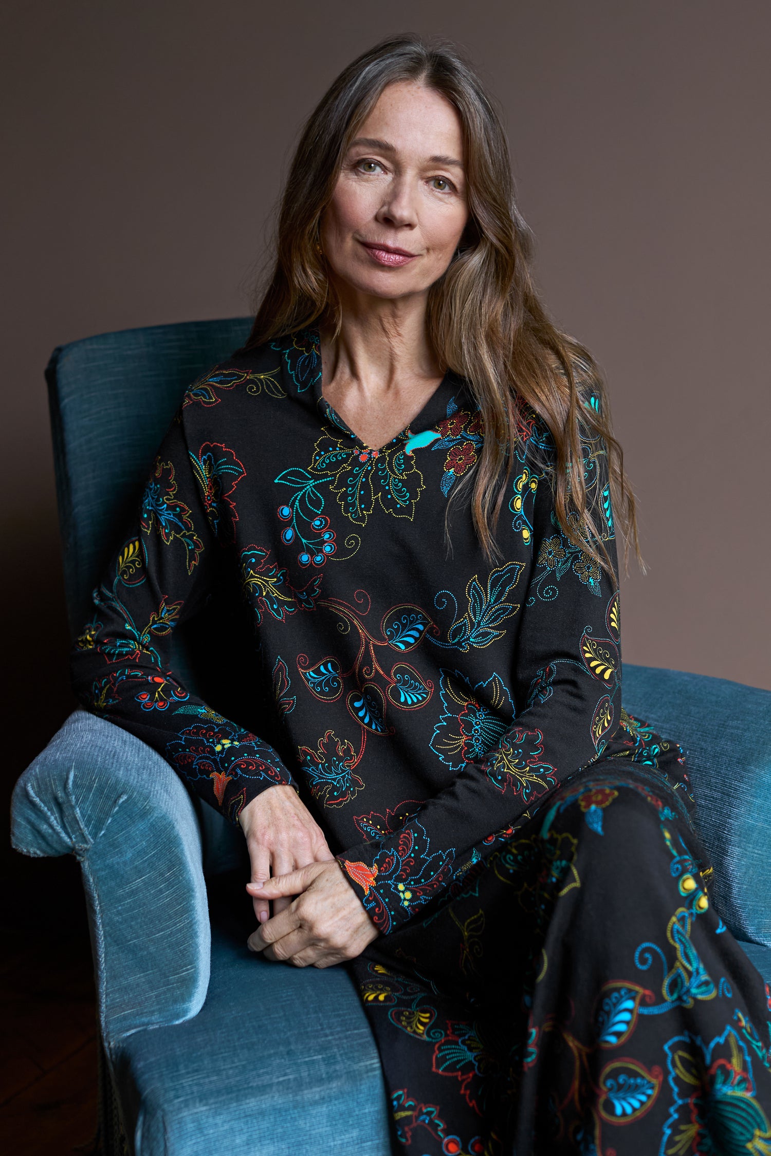 A woman with long hair sits on a blue armchair, wearing the Bright Dotted Floral Jersey Dress, a V-neckline black dress adorned with vibrant floral patterns perfect for day-to-night dressing, against a plain background.