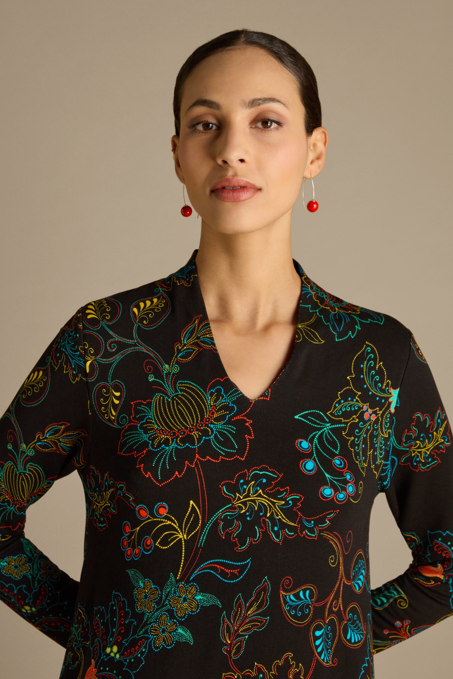 Standing against a neutral background, a woman with red earrings wears the Bright Dotted Floral Jersey Dress. The dress is crafted from warm-handle jersey fabric and showcases a vibrant floral design with a V-neckline, while her hair is tied back.