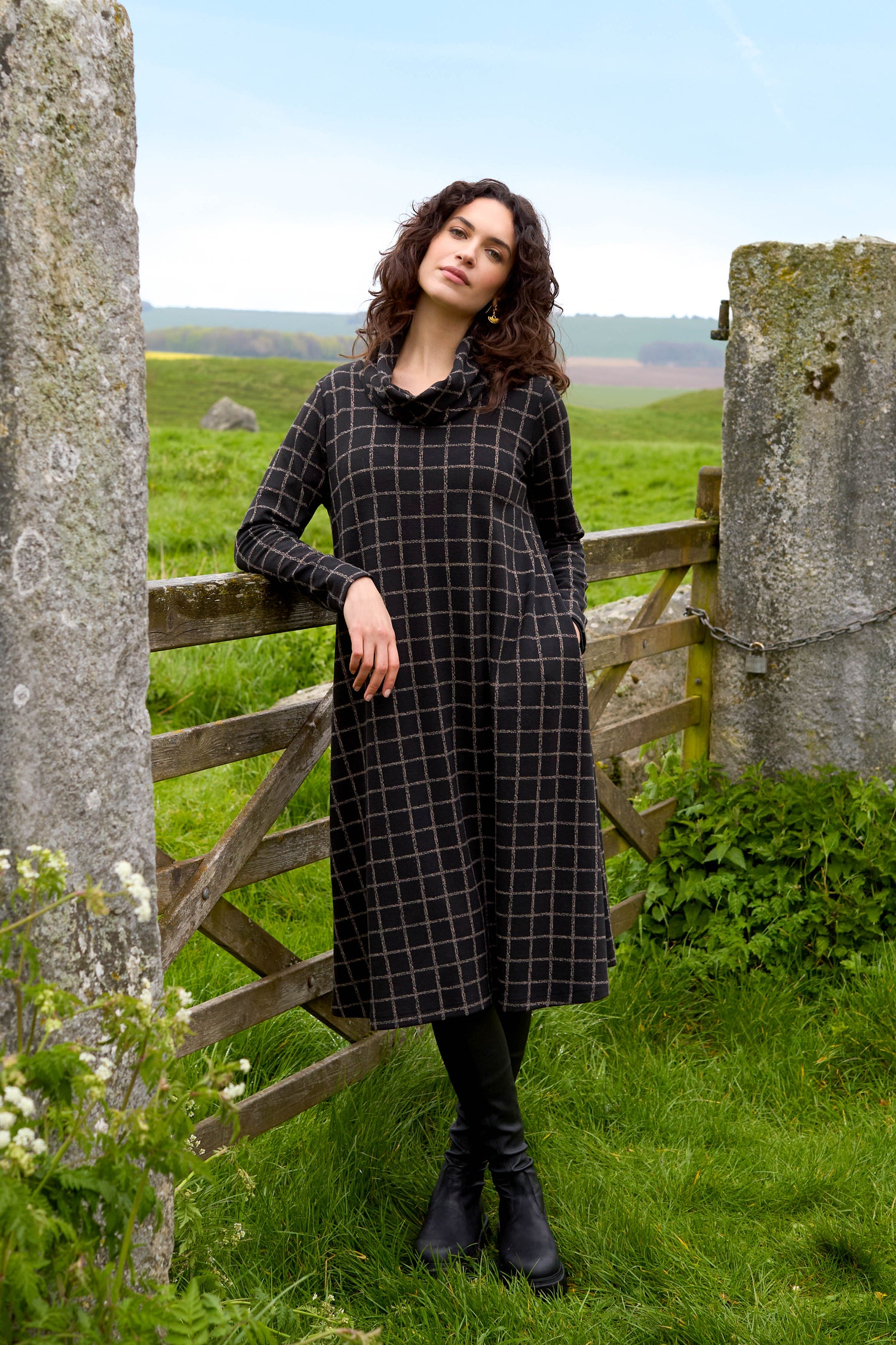 A person wearing a Broken Check Jersey Dress and black boots stands by a wooden gate in a grassy area, with stone pillars and a distant field in the background.