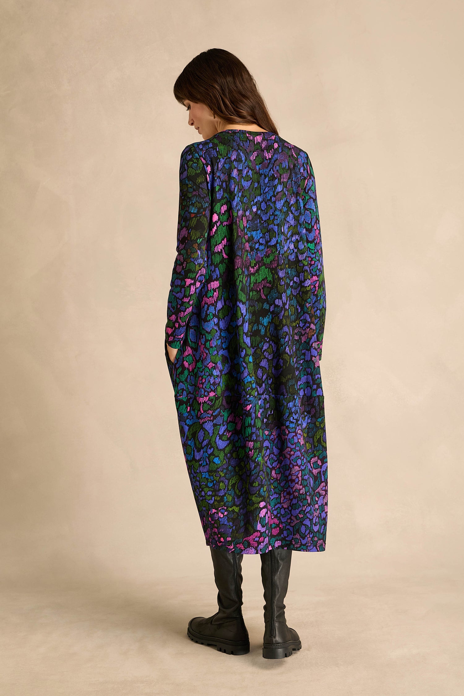 A person stands with their back to the camera, wearing the Winter Garden Jersey Bubble Dress—a loose, long-sleeved style featuring a vibrant blue, purple, and green pattern—paired with black boots against a plain beige background.