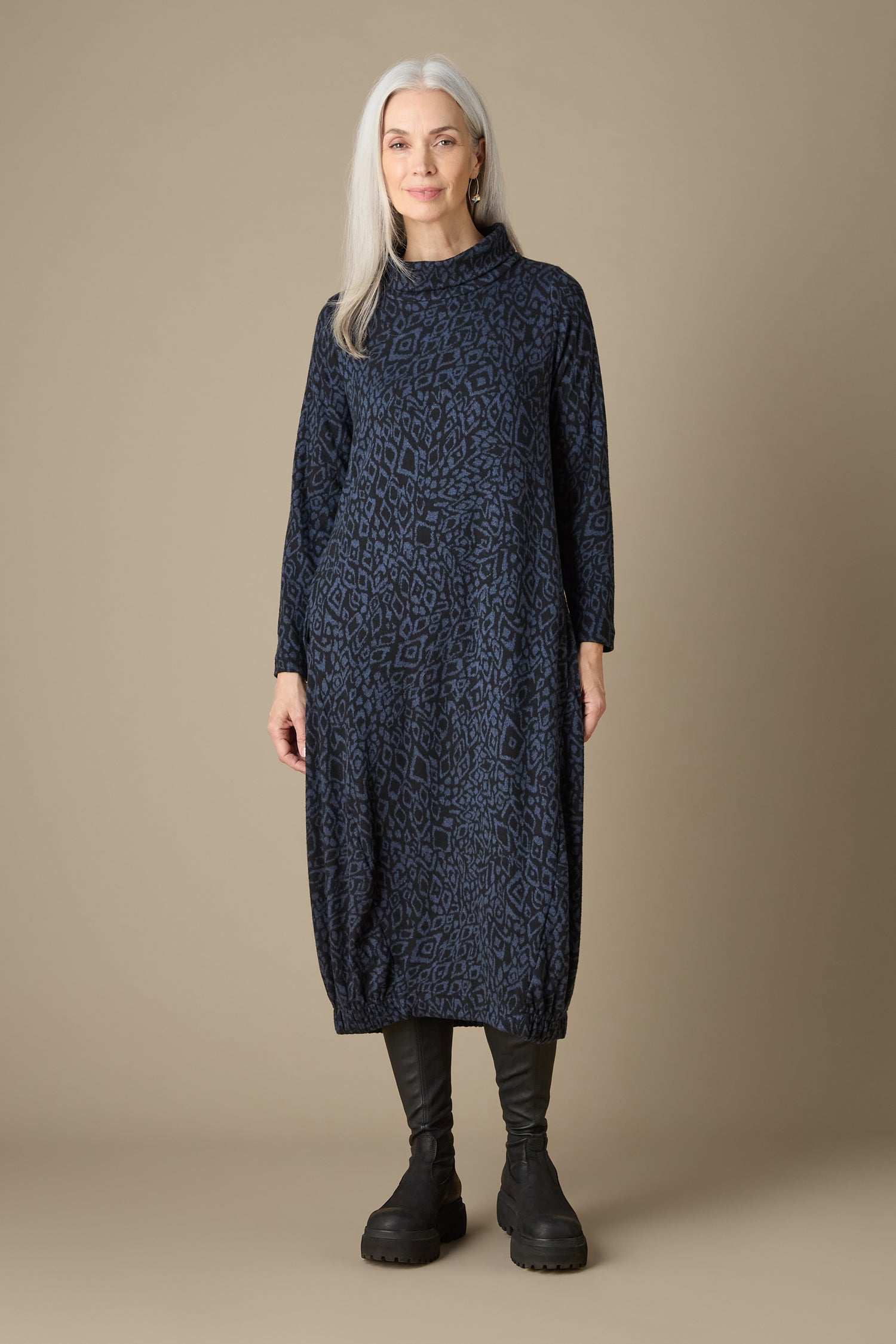 A woman with long gray hair stands against a beige background, wearing an Ikat Soft Jersey Bubble Dress paired with black boots. The distinctive silhouette of her patterned navy dress enhances the artistic quality of the scene.
