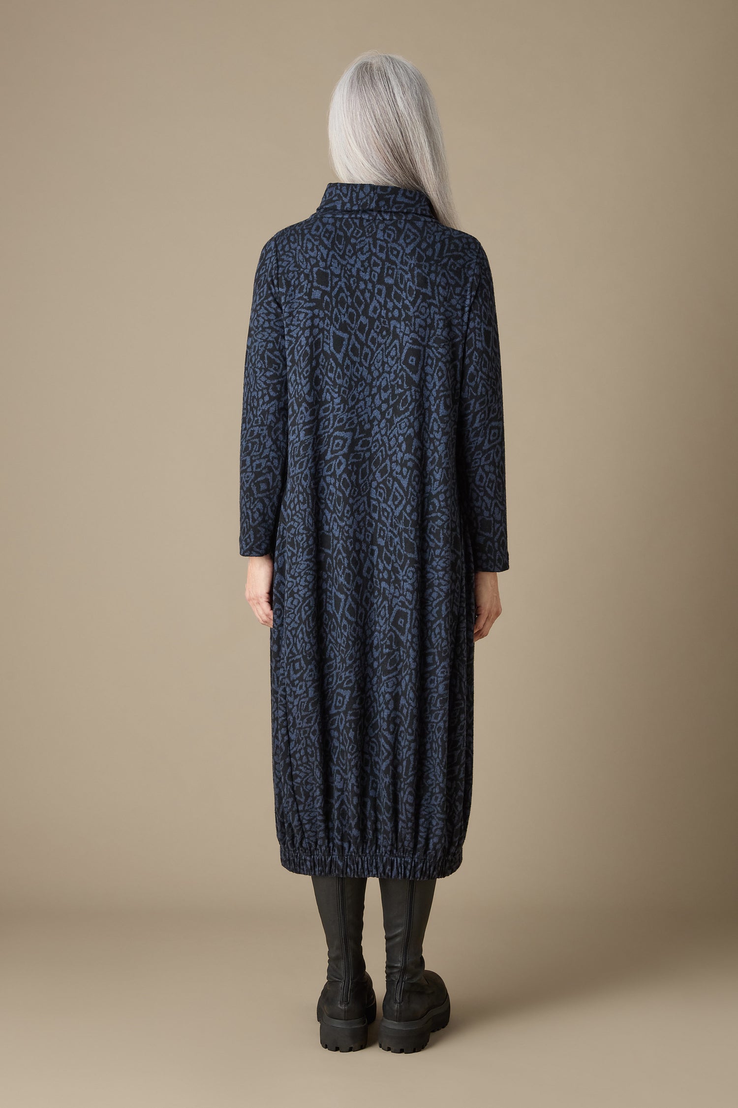 A person with long gray hair is wearing a long, dark blue coat featuring an abstract print, paired with black boots. They are standing against a beige background, shown from the back in what resembles the Ikat Soft Jersey Bubble Dress design.