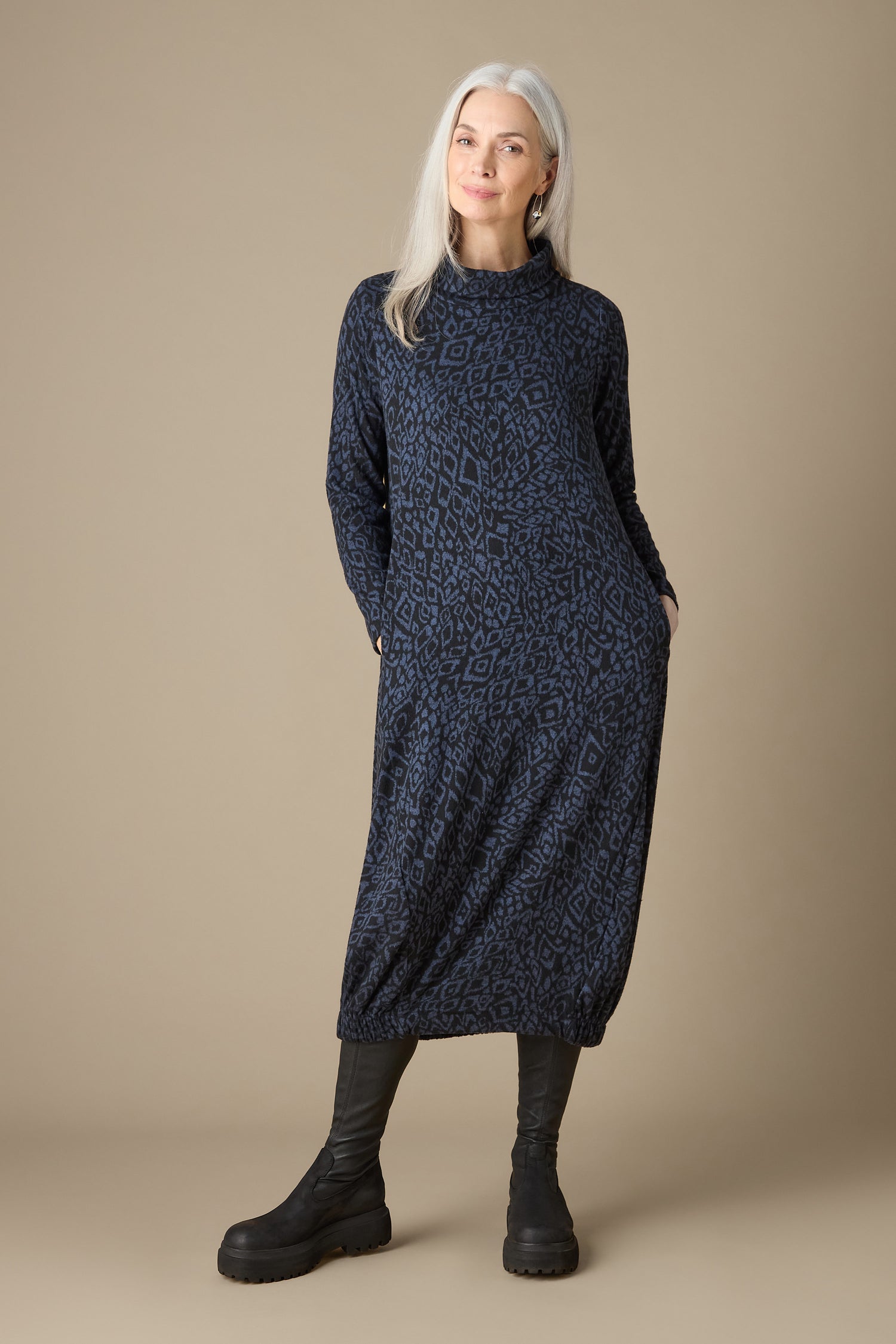A woman with long gray hair poses in front of a beige background, dressed in the Ikat Soft Jersey Bubble Dress. This dress, with its distinctive bubble silhouette and dark abstract pattern, complements her black boots beautifully.