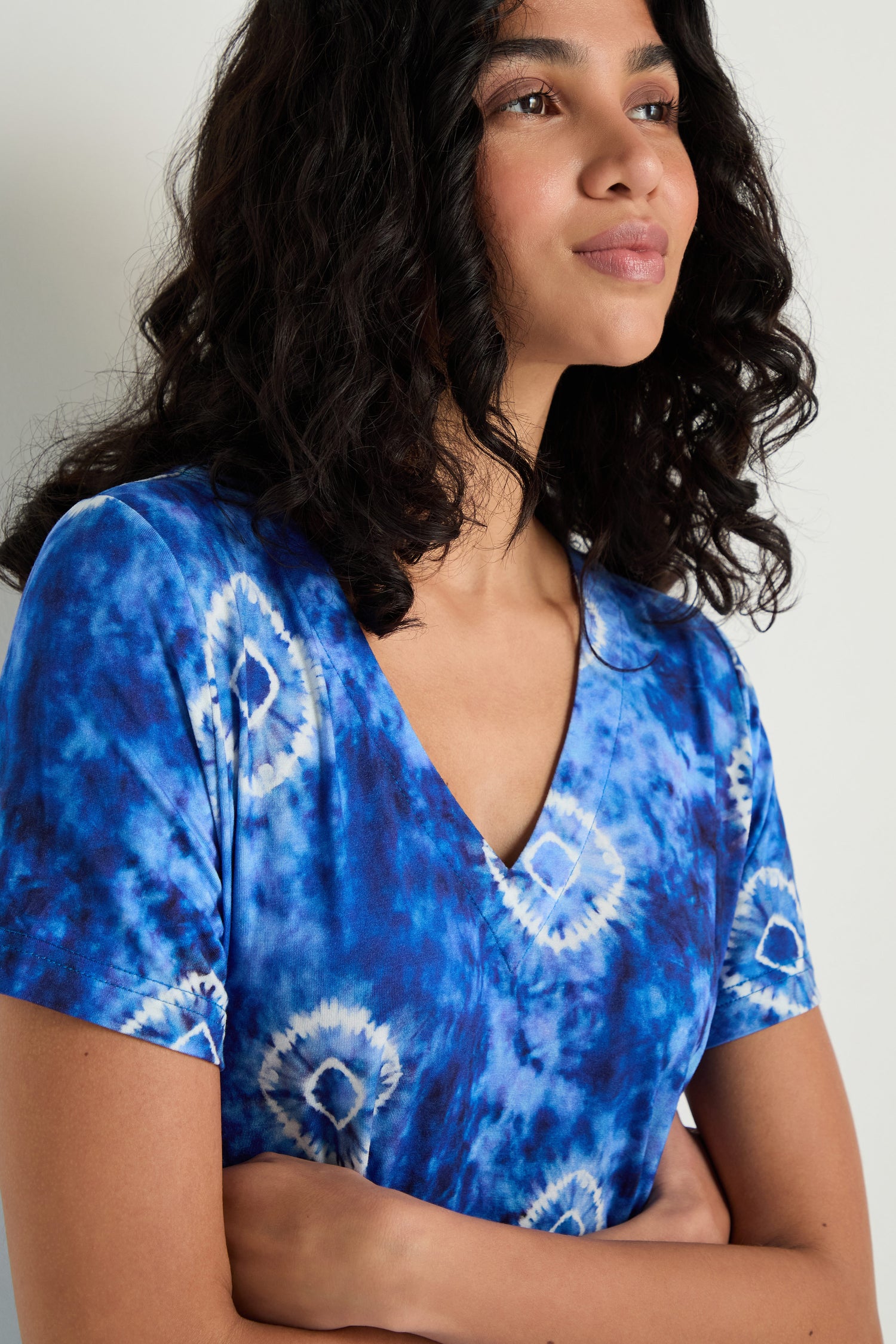 A woman with wavy dark hair wears the Tie Dye Circles Jersey Bubble Dress and stands with her arms crossed, looking slightly to the side.