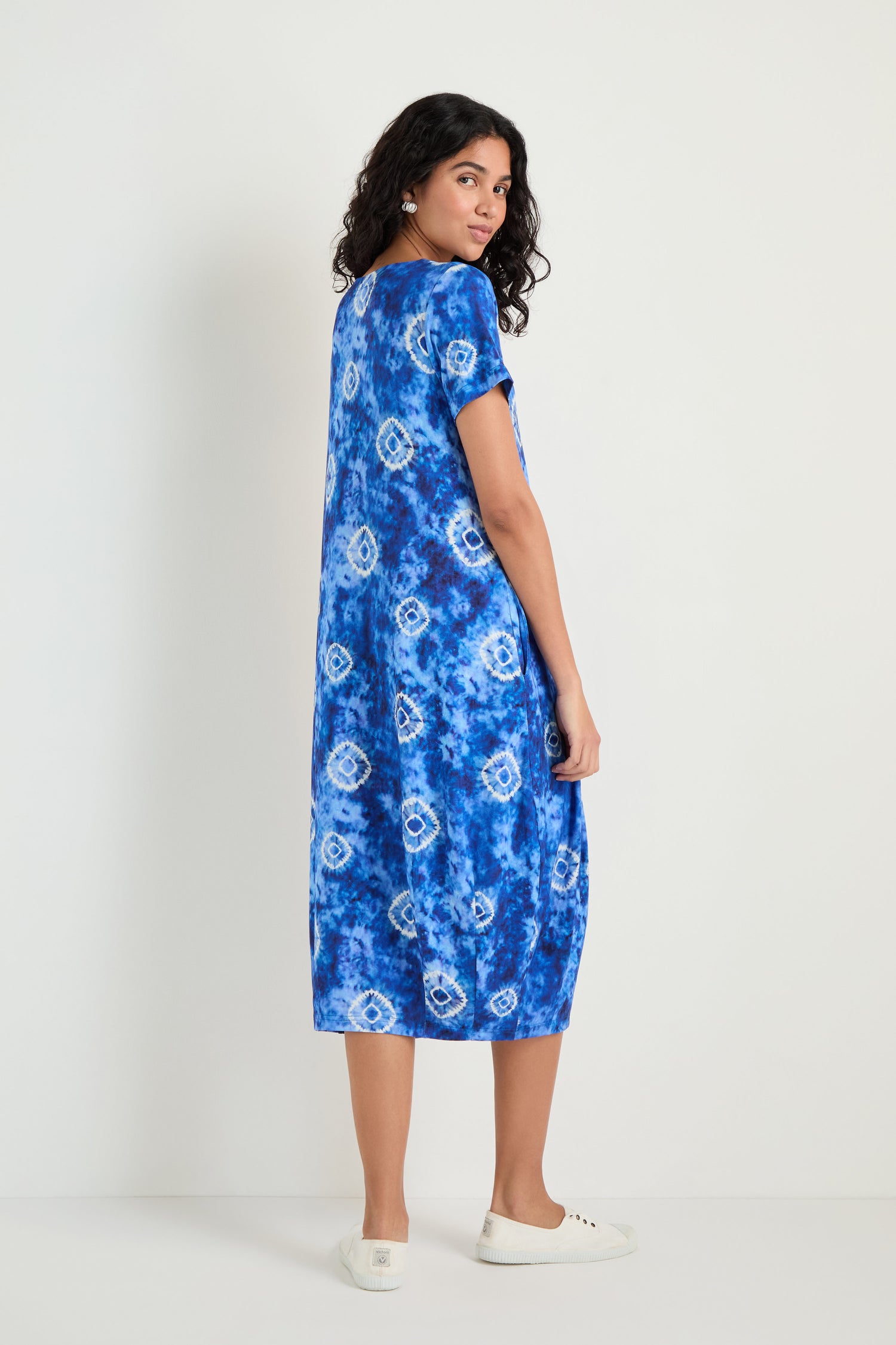 A woman poses against a plain background in the Tie Dye Circles Jersey Bubble Dress with short sleeves and white sneakers, looking over her shoulder.