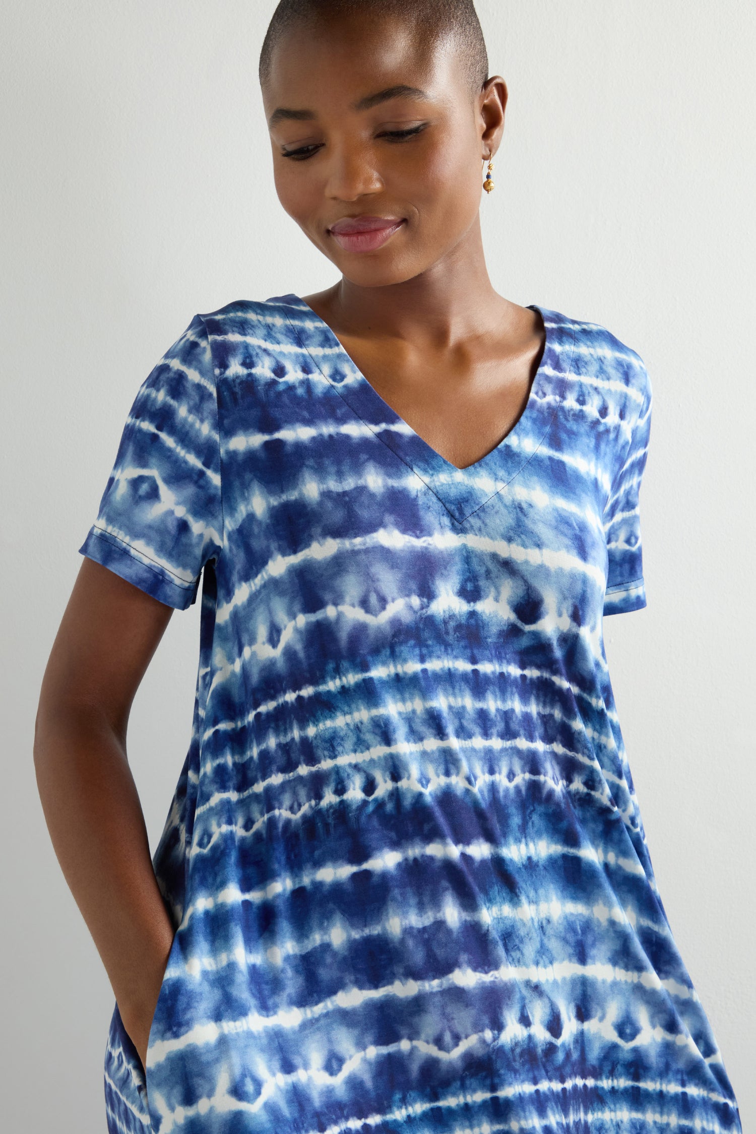 A person wearing the Tie Dye Jersey Bubble Dress with short sleeves stands against a light background, hands in pockets, and smiles downwards. Their relaxed silhouette radiates ease and casual elegance.