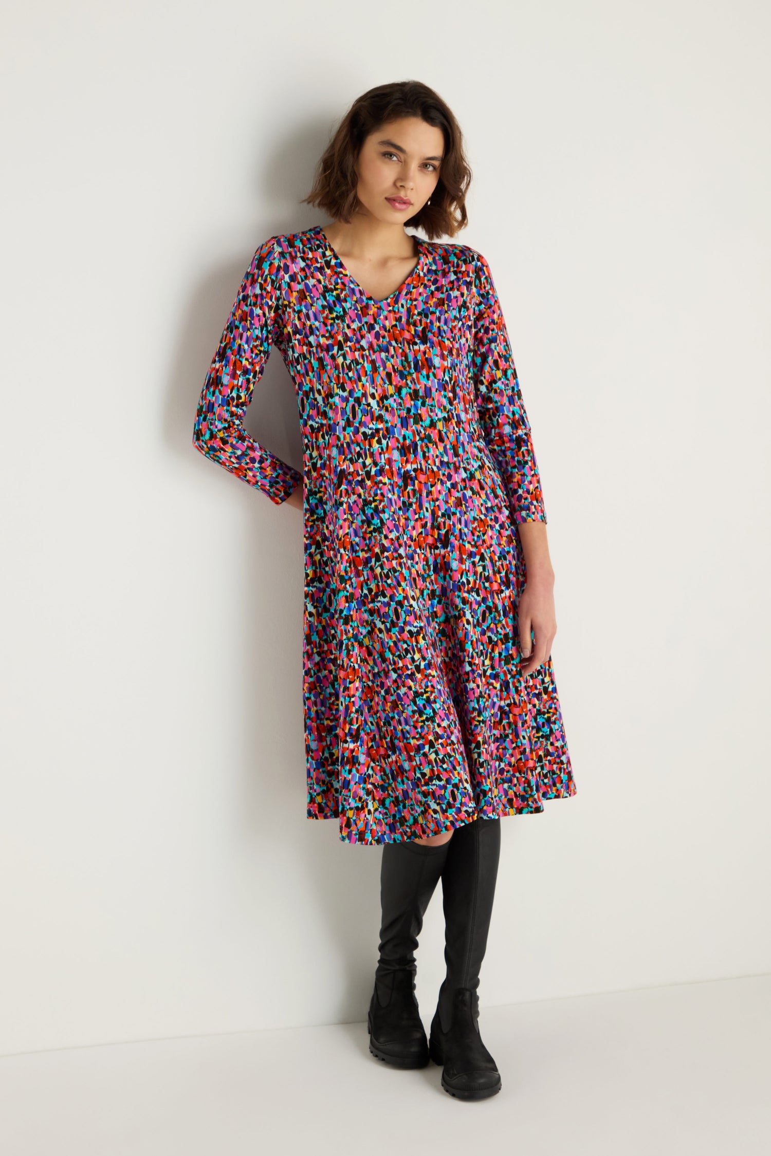A woman stands against a white wall wearing the Painted Spots Jersey Flared Dress, featuring a colorful abstract print, paired with black leggings and black shoes.