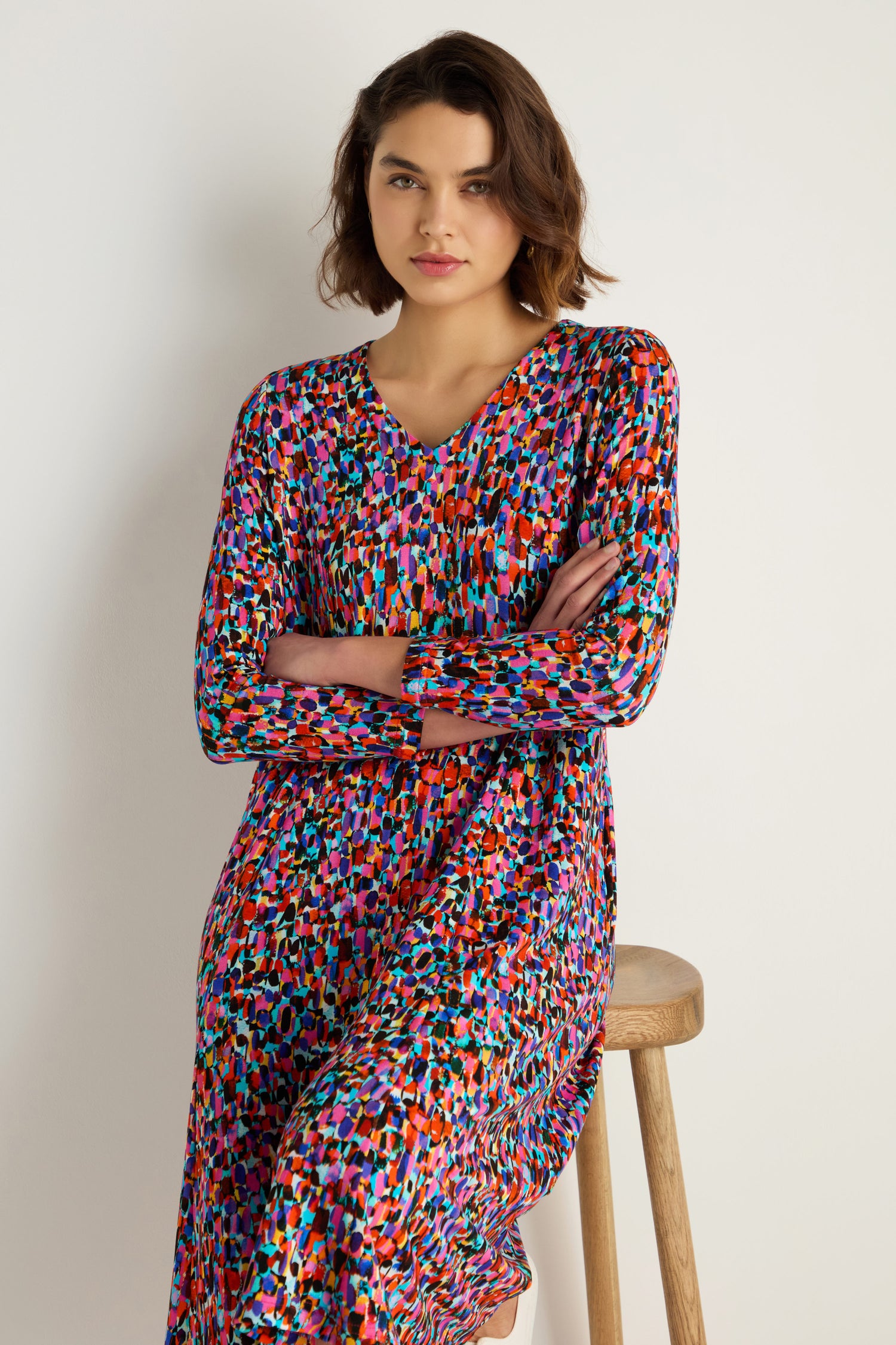 A woman with short brown hair sits on a wooden stool against a plain white background, wearing the Painted Spots Jersey Flared Dress, a long-sleeved, midi-length piece featuring colorful painted spots.