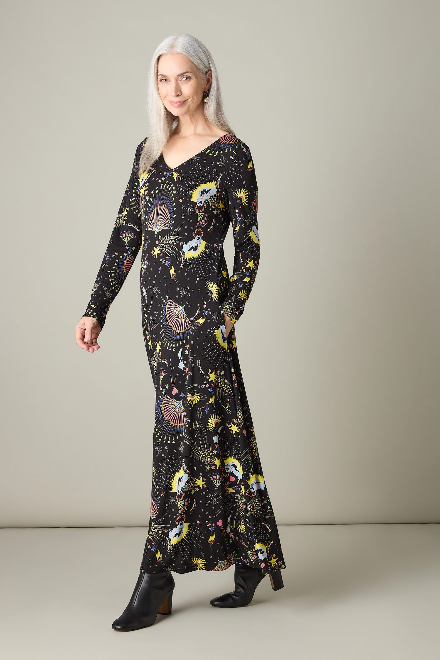 A woman with long grey hair is wearing the Celestial Dreams Jersey A Line Maxi Dress, showcasing an A-line silhouette embellished with a vibrant celestial pattern. She complements it flawlessly with black ankle boots while standing confidently against a plain background.