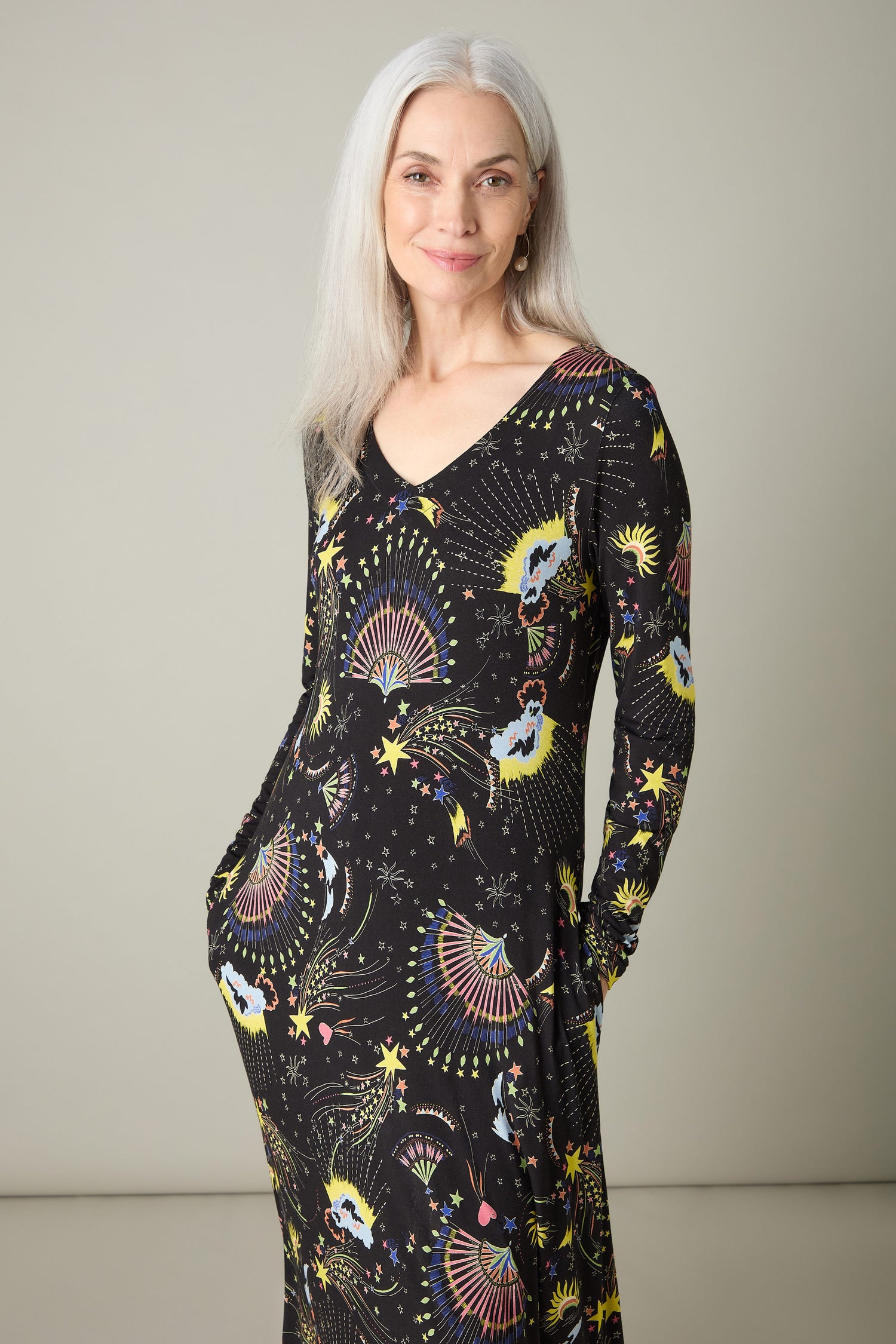 A person with long gray hair dons a black dress, accentuated by a vibrant celestial print in an abstract design. They stand against a plain background, smiling with hands in pockets—an ideal representation of the Celestial Dreams Jersey A Line Maxi Dress's charm.