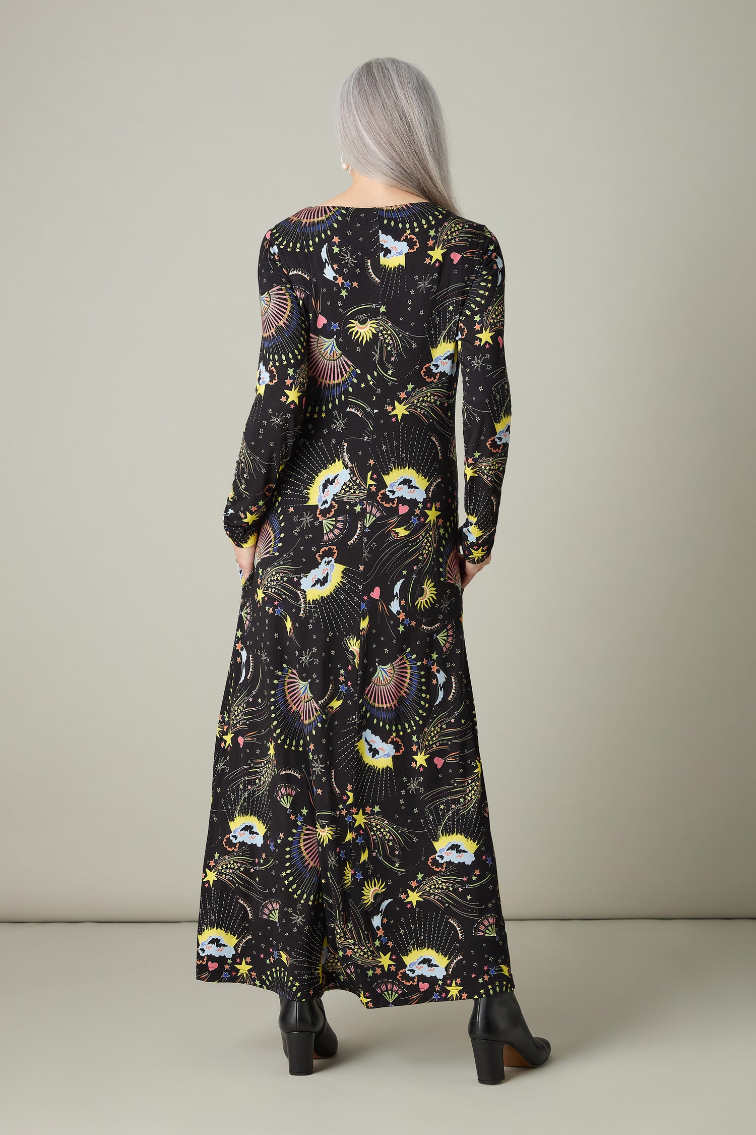 A person with long gray hair stands facing away, wearing the Celestial Dreams Jersey A Line Maxi Dress adorned with a celestial print and paired with black heels against a plain light background.