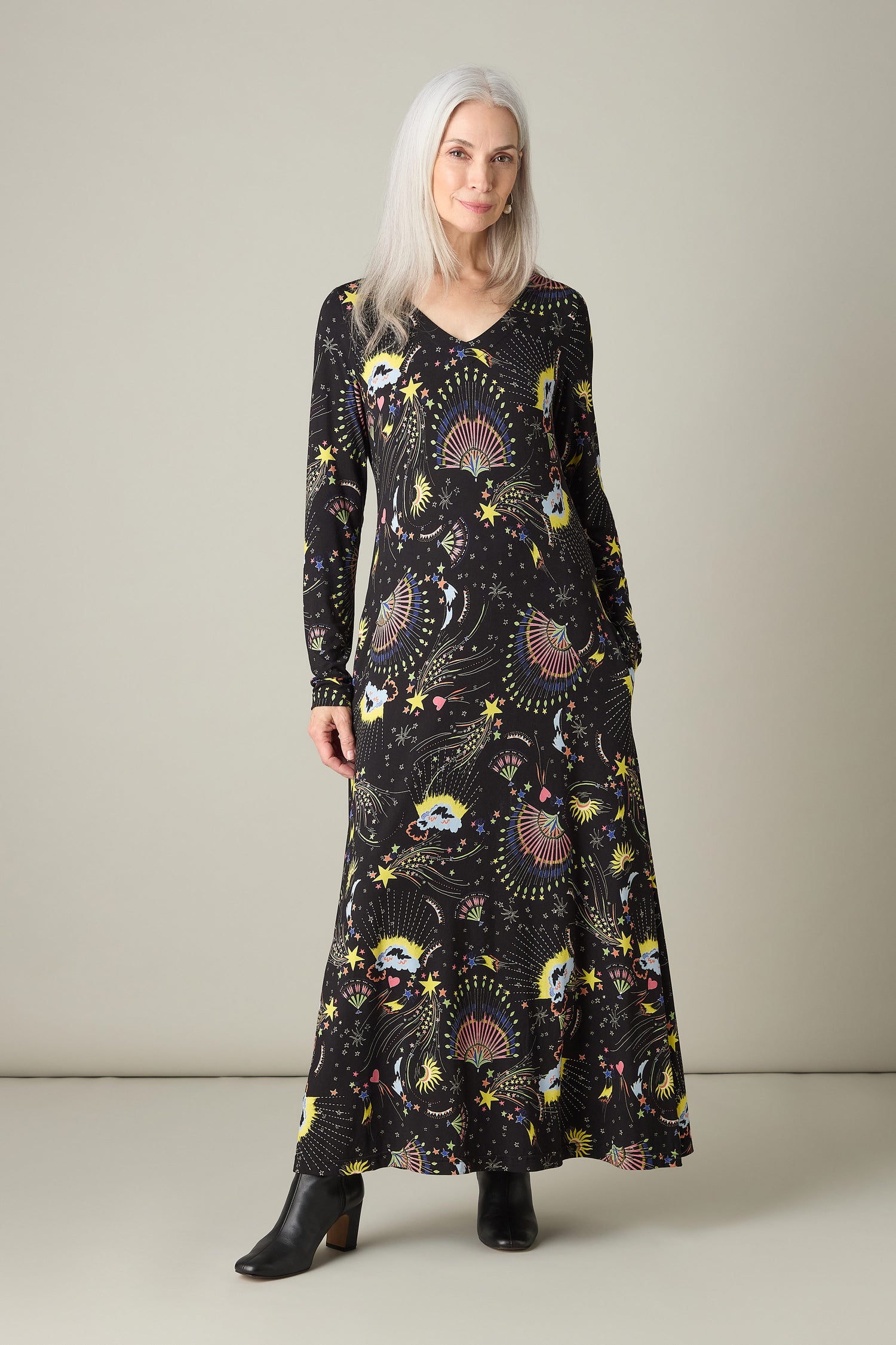 A woman with long gray hair stands against a plain background wearing the Celestial Dreams Jersey A-Line Maxi Dress, featuring a striking celestial print. She completes her look with stylish black ankle boots.