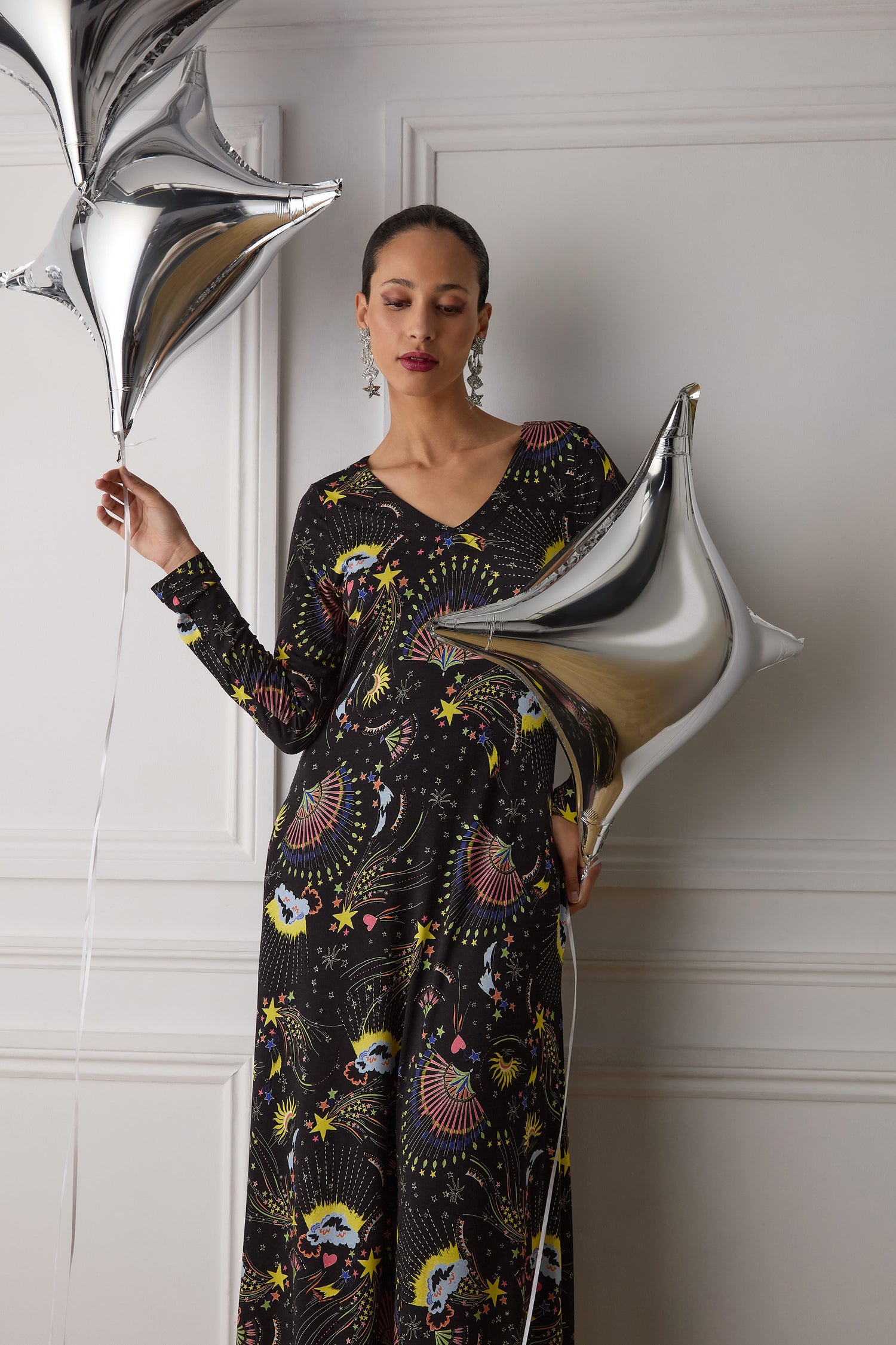 A person wearing the Celestial Dreams Jersey A Line Maxi Dress holds two silver star-shaped balloons against a white paneled wall.