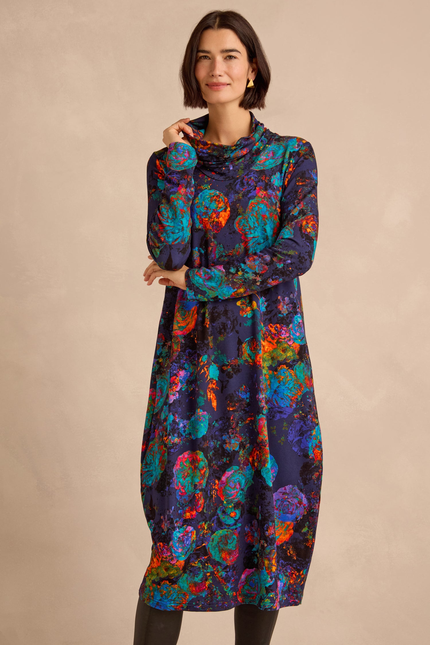 Wearing the Midnight Floral Jersey Bubble Dress, a woman stands in a long-sleeve, high-neck piece featuring an abstract floral pattern set against a deep background and finished with an elegant midi-length bubble hem.