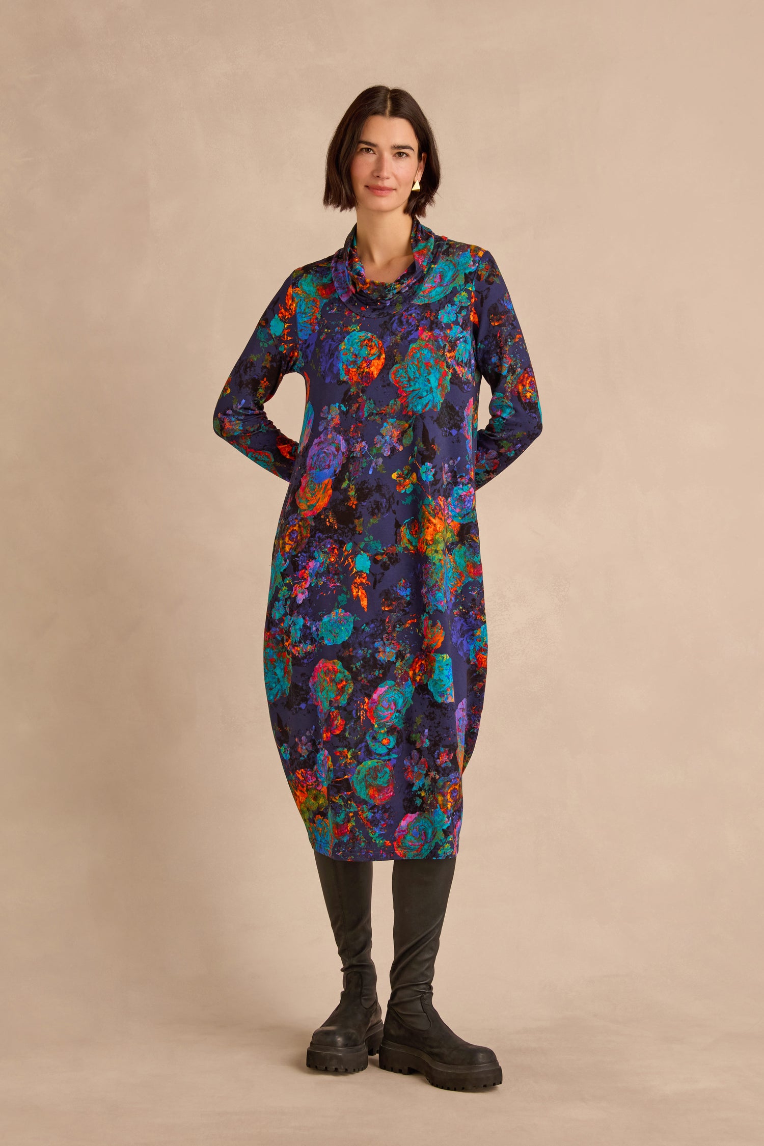 A woman stands against a neutral background, wearing the Midnight Floral Jersey Bubble Dress in a midi length with a colorful abstract pattern, paired with black boots.