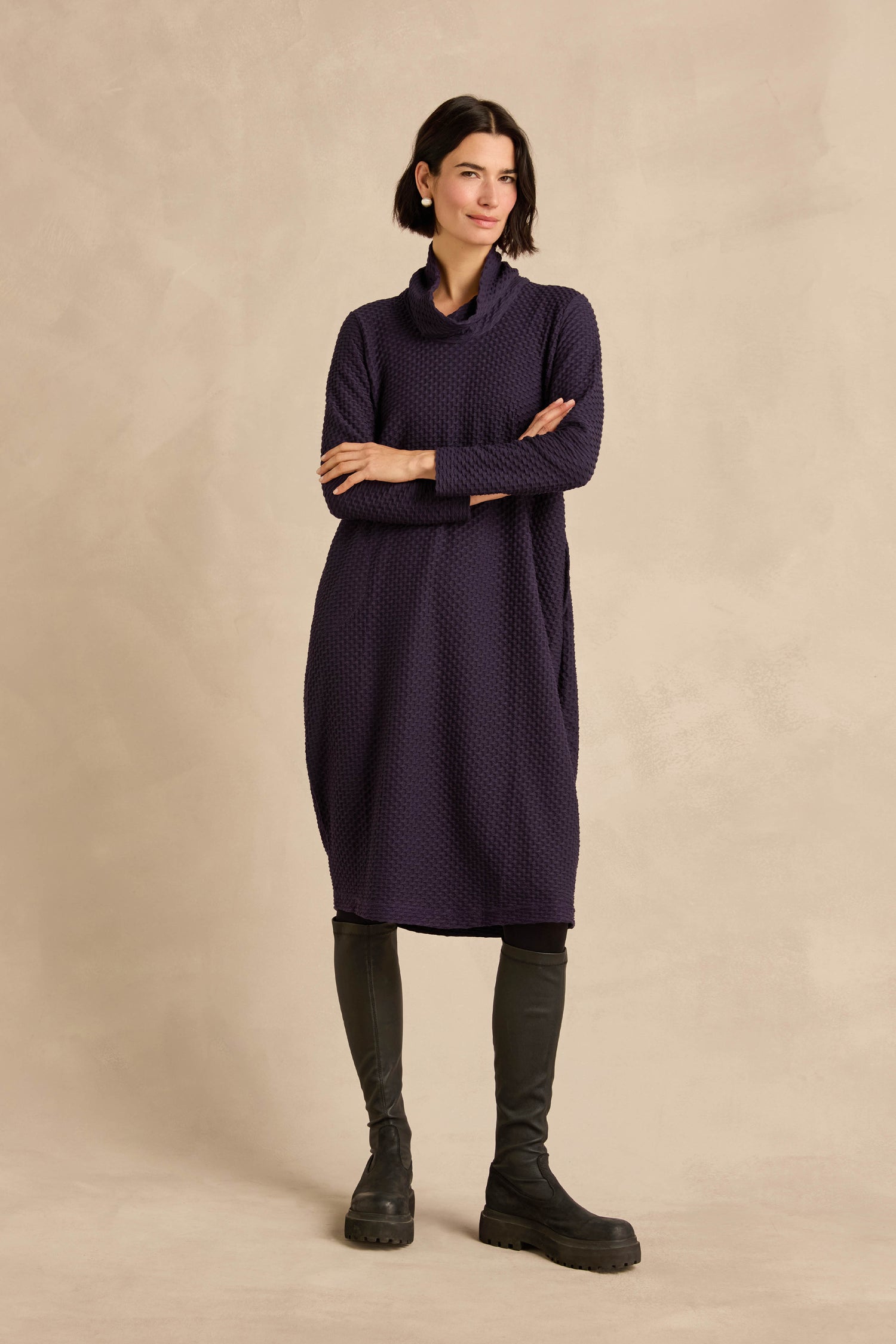 A woman stands indoors against a beige backdrop, arms crossed and looking slightly to the side, wearing the Damson Waffle Textured Jersey Bubble Dress with long sleeves and black knee-high boots.