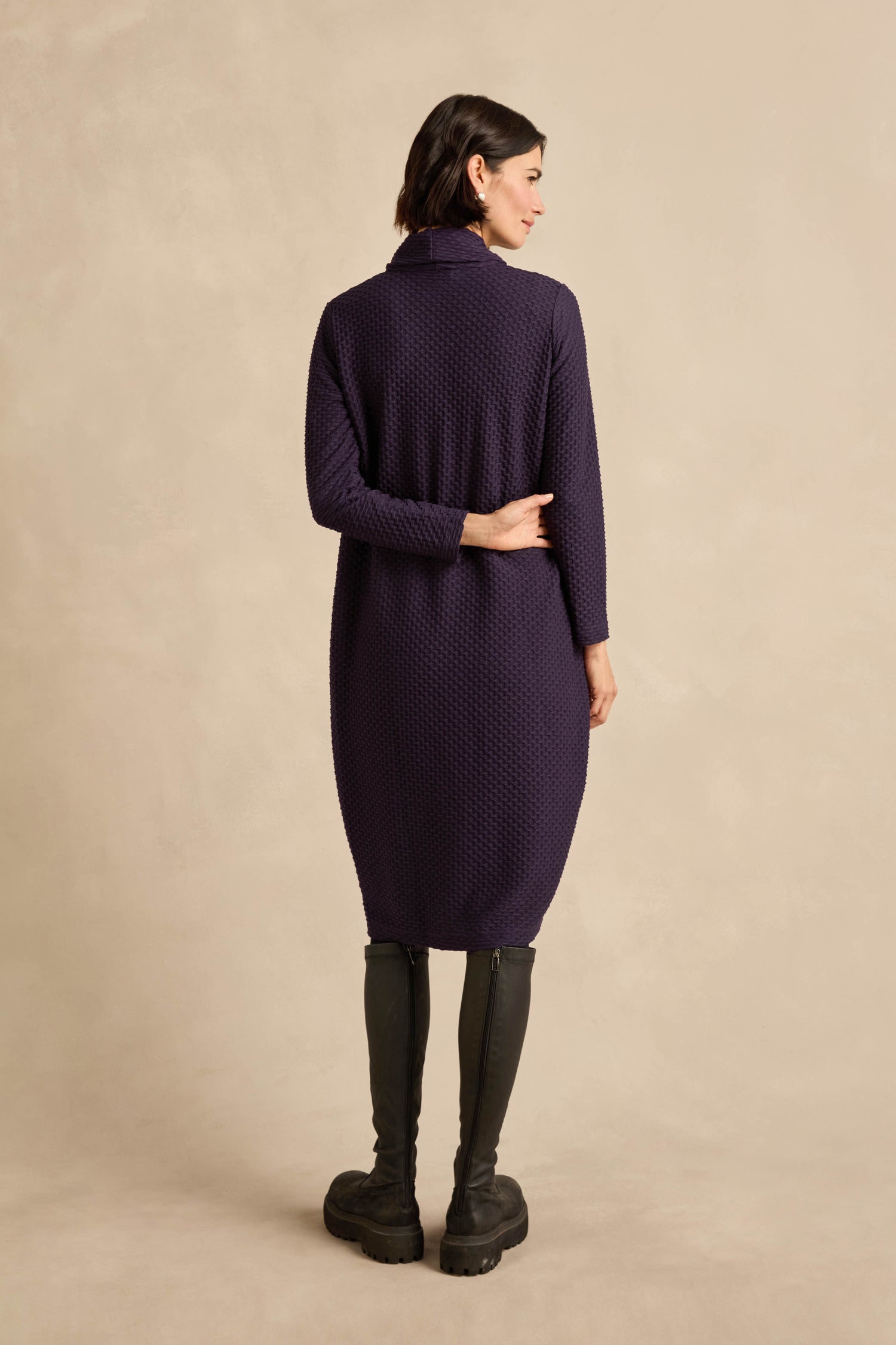 A woman stands with her back to the camera, dressed in the Damson Waffle Textured Jersey Bubble Dress and black knee-high boots, set against a plain beige background.