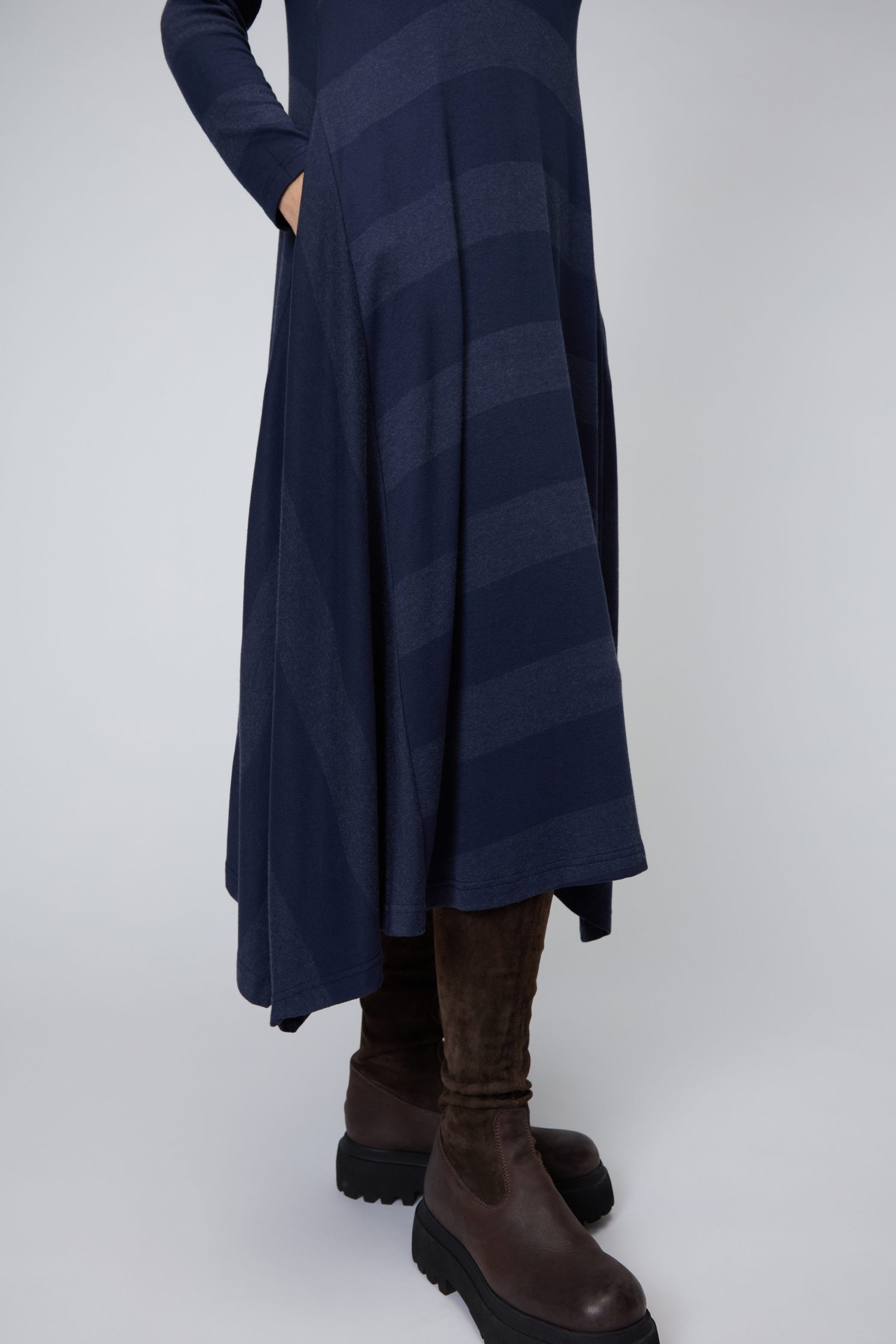 A person wears the Soft Viscose Jersey Stripe Dress—navy blue, long-sleeved, with stripes and an asymmetric hem—paired with brown knee-high boots, standing against a plain background.