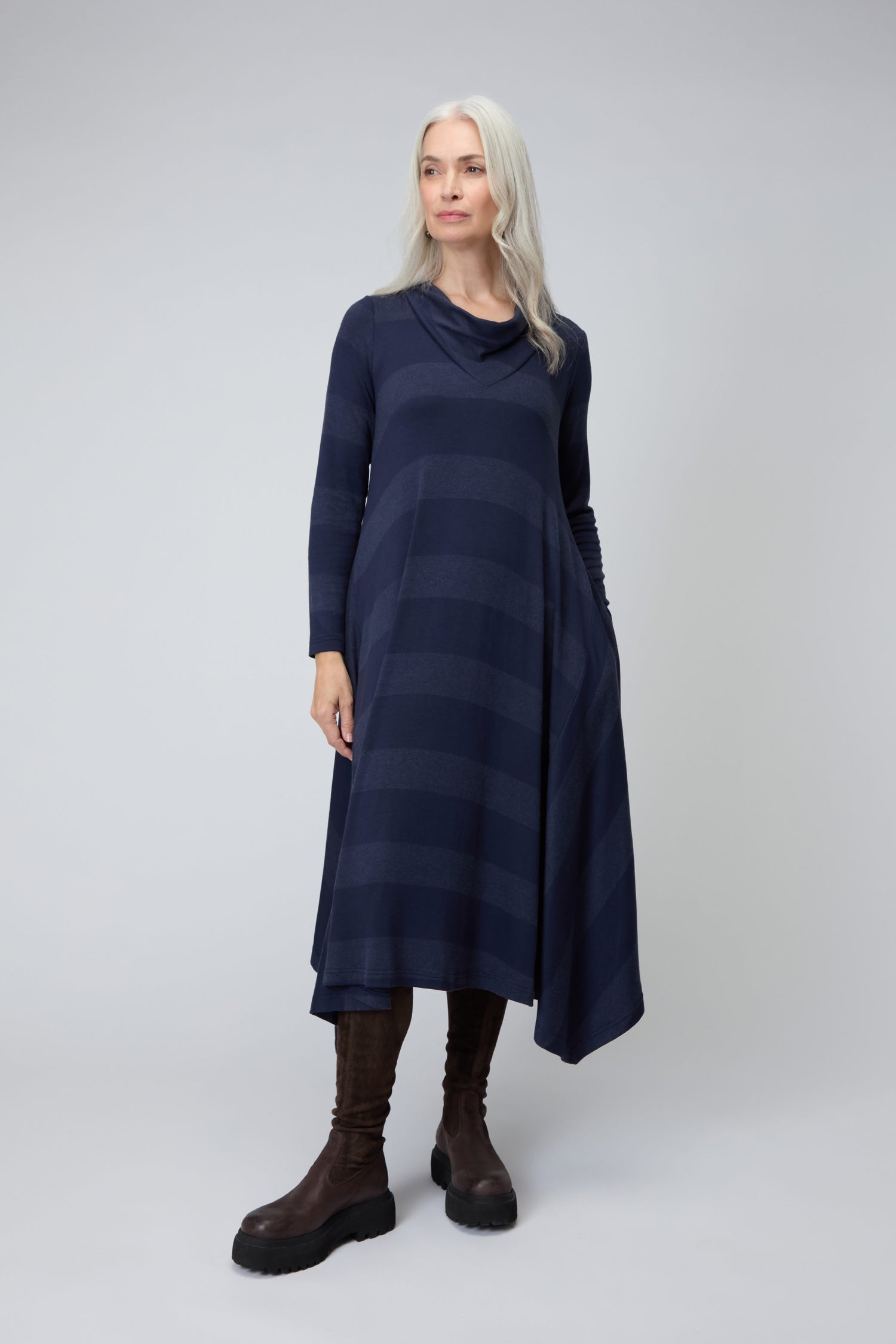 A woman with long gray hair models the Soft Viscose Jersey Stripe Dress in dark blue, featuring subtle stripes, an asymmetric hem, and long sleeves. She pairs it with brown knee-high boots against a plain light background.