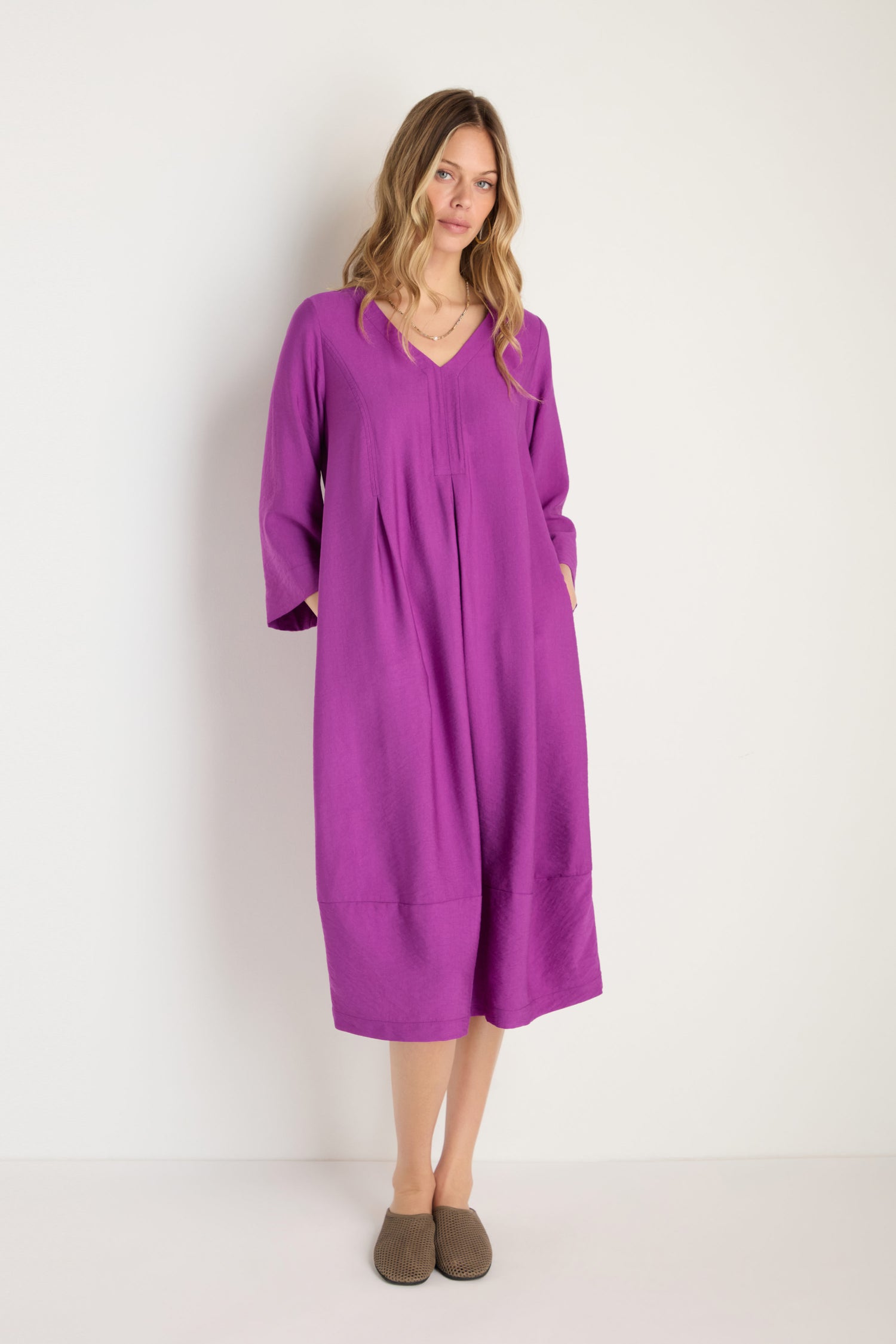 A woman stands against a white wall wearing a loose-fitting Textured Viscose Bubble Dress in orchid purple with long sleeves, paired with brown slippers.