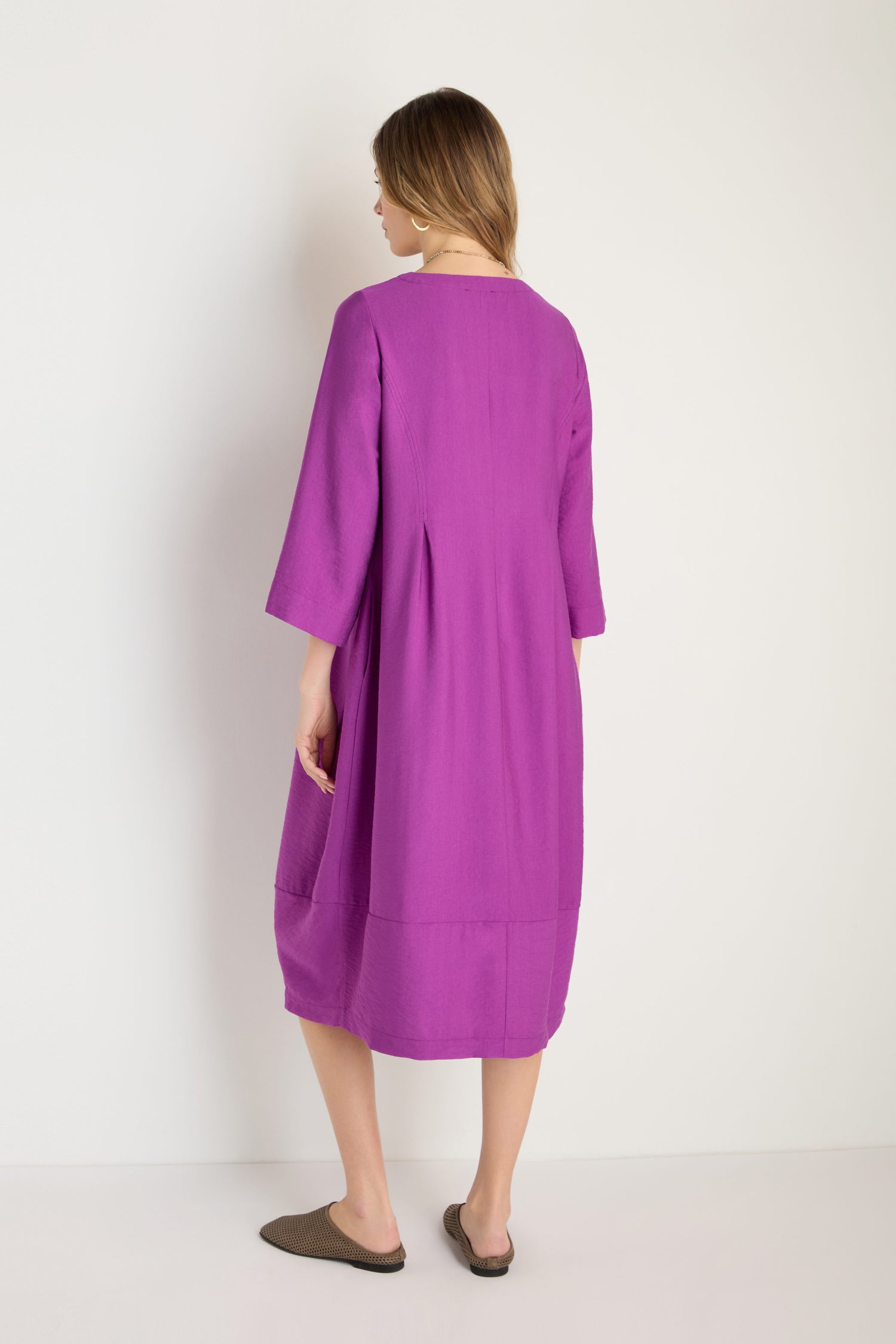 A woman stands with her back to the camera, wearing the Textured Viscose Bubble Dress in orchid purple—a loose-fitting, knee-length style with three-quarter sleeves—against a plain white background.