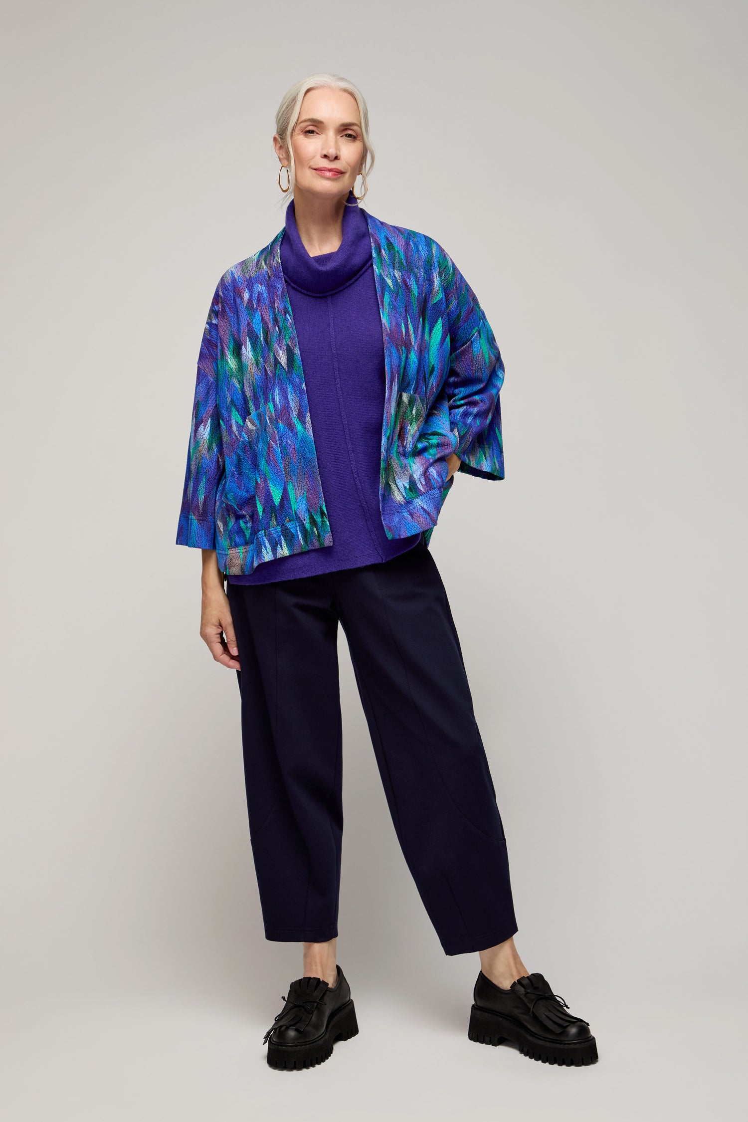 A woman with gray hair in a low ponytail wears the Abstract Feather Jersey Jacket over a blue turtleneck, dark pants, and black shoes while standing against a plain background.