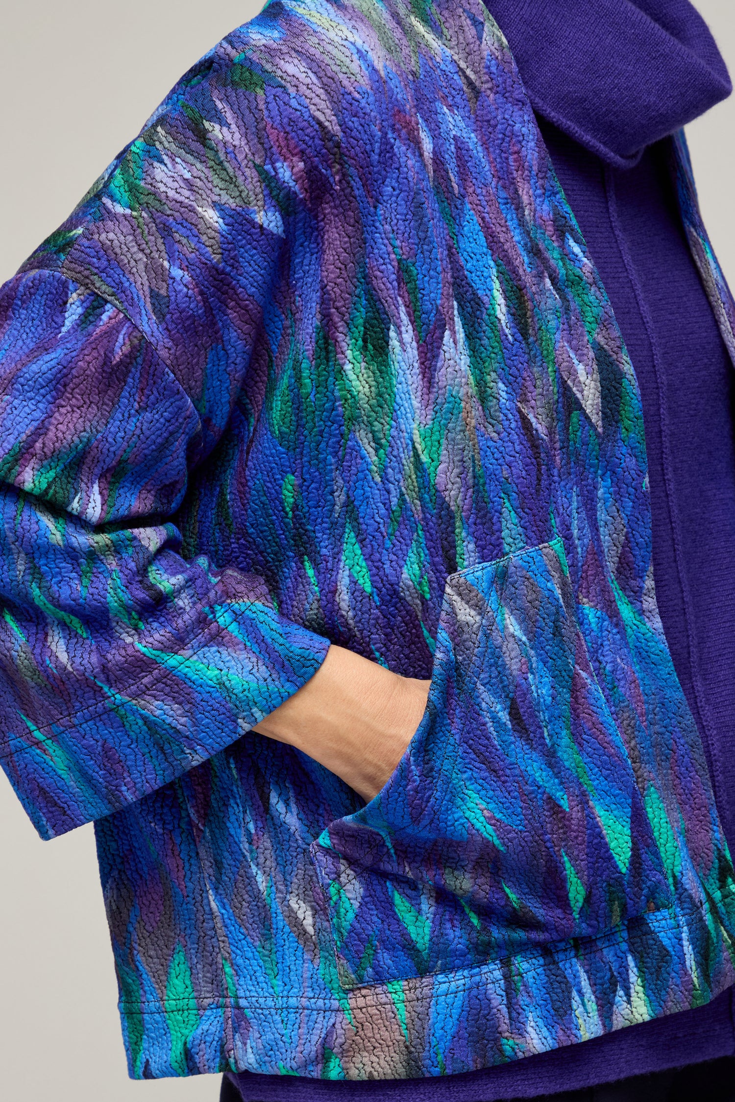 A person wears the Abstract Feather Jersey Jacket, a cropped boxy style in textured blue and purple, with one hand in the pocket over a solid purple top—a bold, modern layering piece.