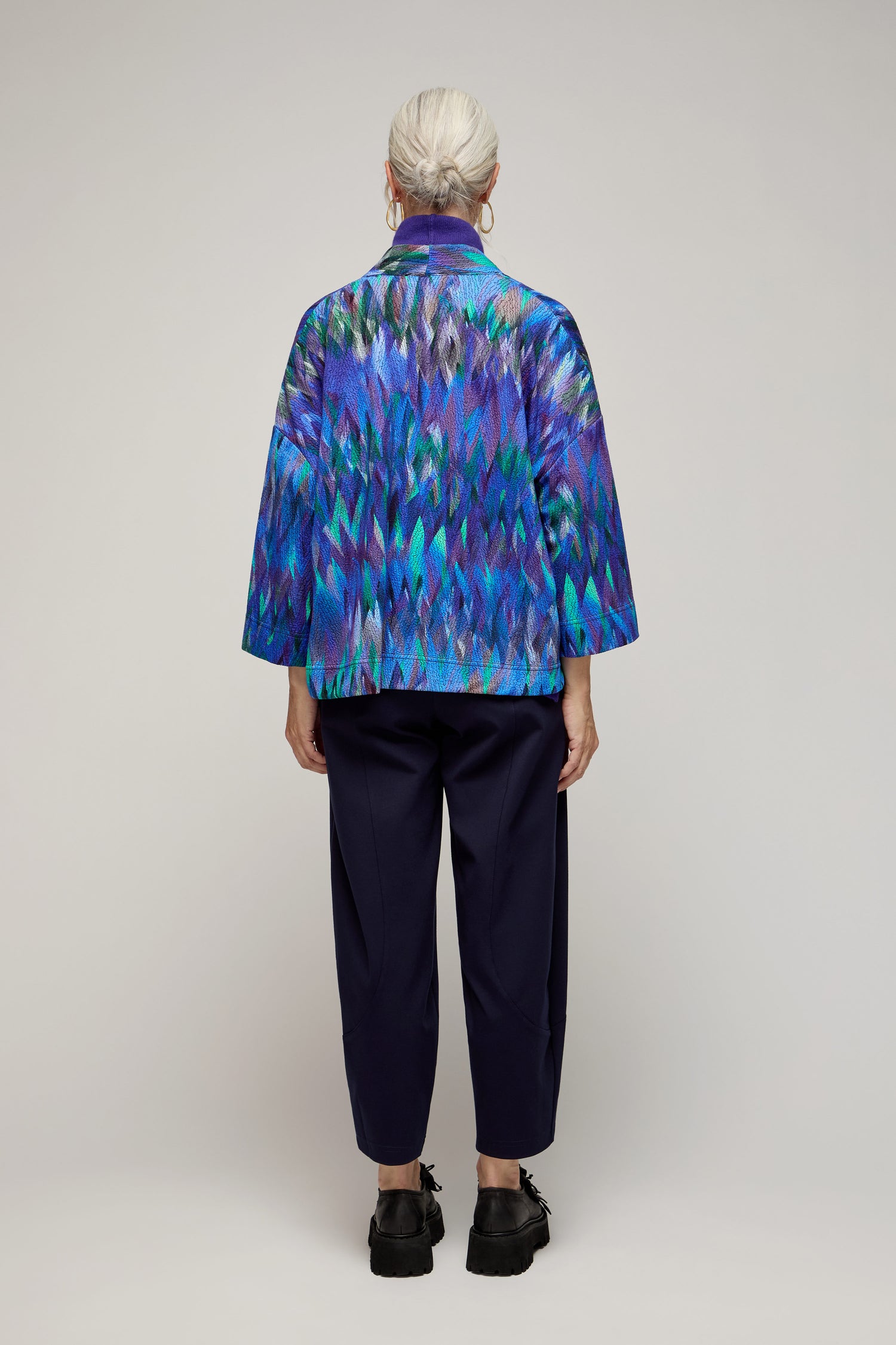 An older person with gray hair stands facing away, wearing an Abstract Feather Jersey Jacket in blue and purple patterns, paired with dark pants and black shoes against a plain light background.