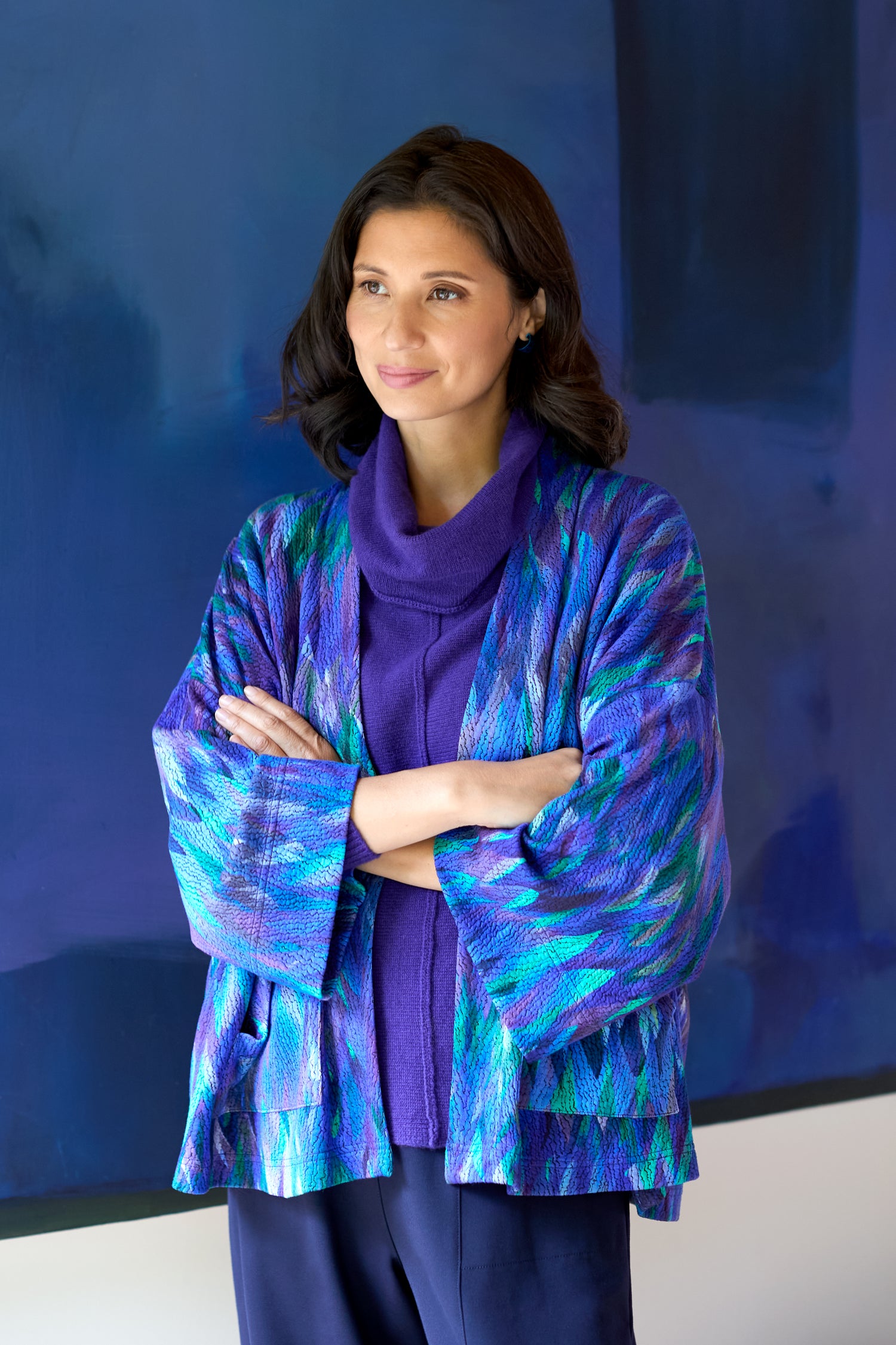 A woman with dark hair stands arms crossed, wearing the Abstract Feather Jersey Jacket—a blue and purple cropped boxy layer—ideal for modern styling, in front of a large abstract blue painting.