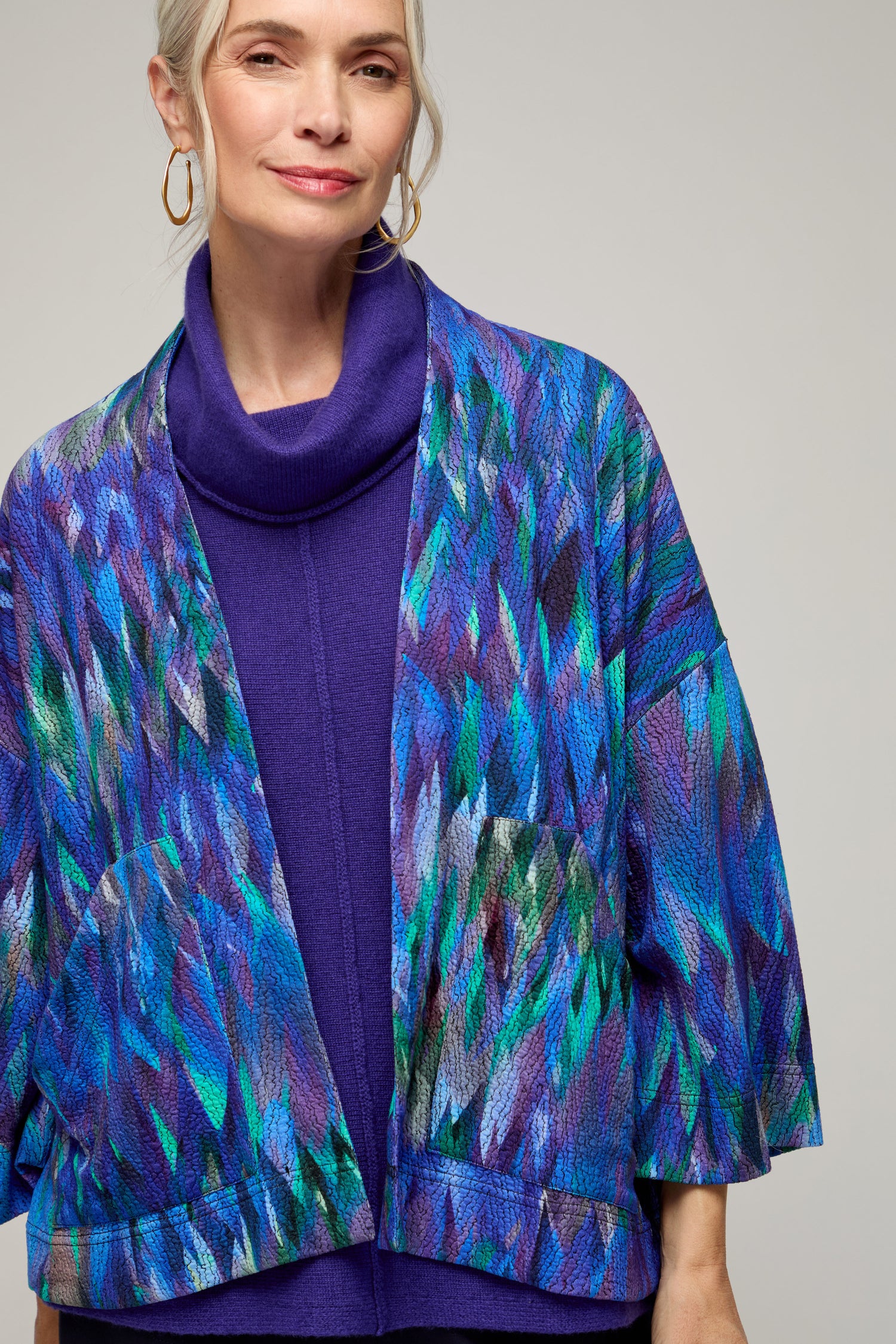 A woman with gray hair stands against a plain background, wearing a purple turtleneck and the Abstract Feather Jersey Jacket featuring a blue, green, and purple feather print.