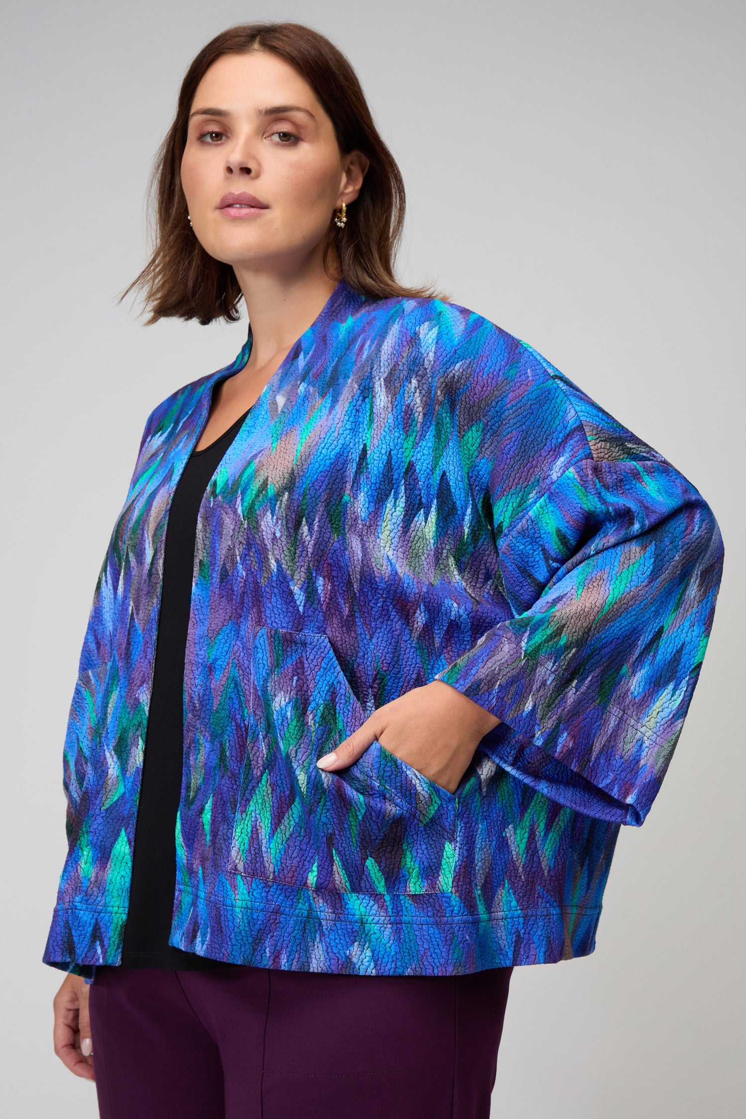 A woman stands with one hand in her pocket, wearing the Abstract Feather Jersey Jacket—featuring blue and purple patterns—over a black top and purple pants, set against a neutral background.