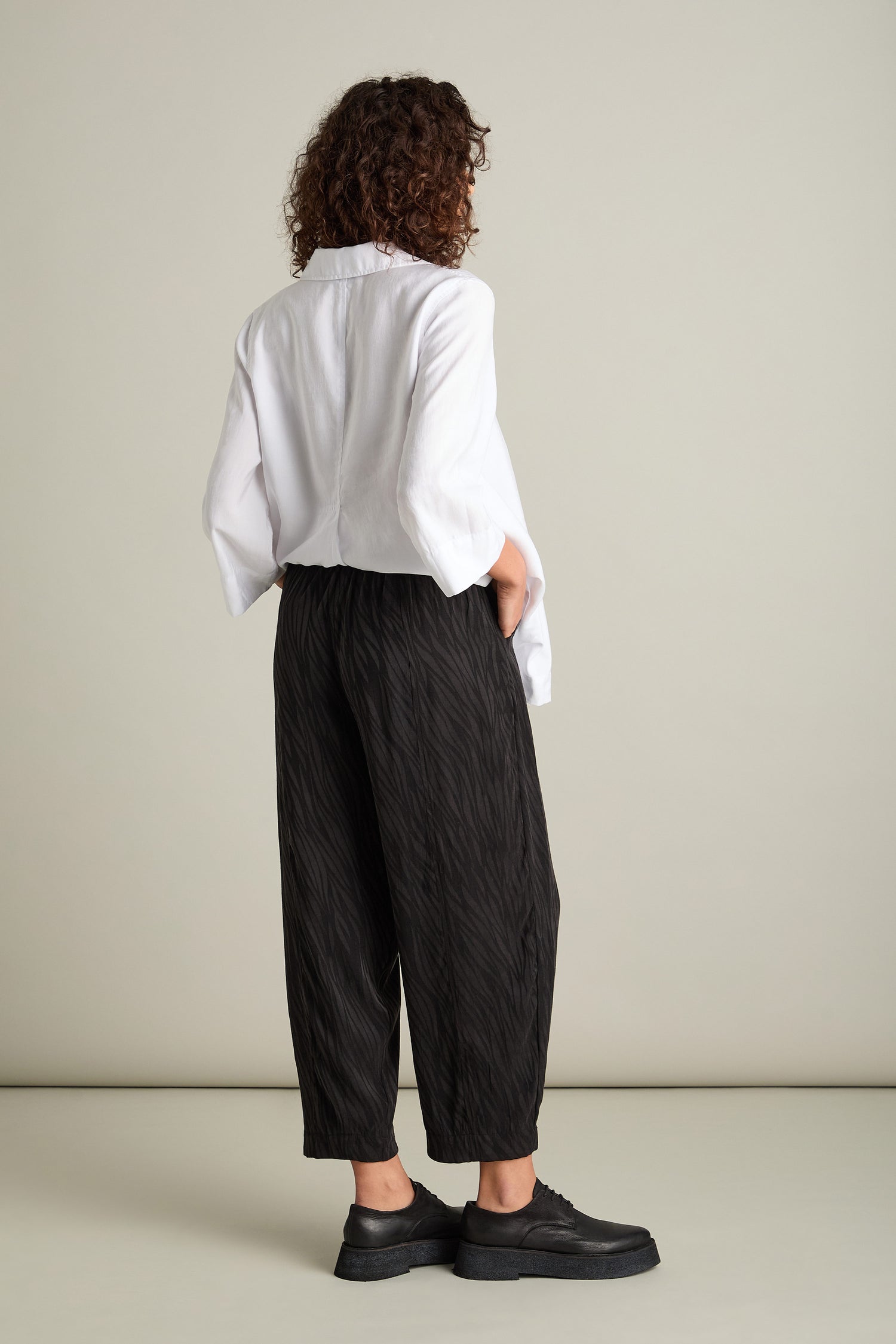 A person with curly hair stands facing away, dressed in a white long-sleeve shirt made from silky-feel fabric and black Abstract Lines Jacquard Bubble Trousers paired with black shoes. Their hands are tucked into their pockets against a plain background, exuding a contemporary look.