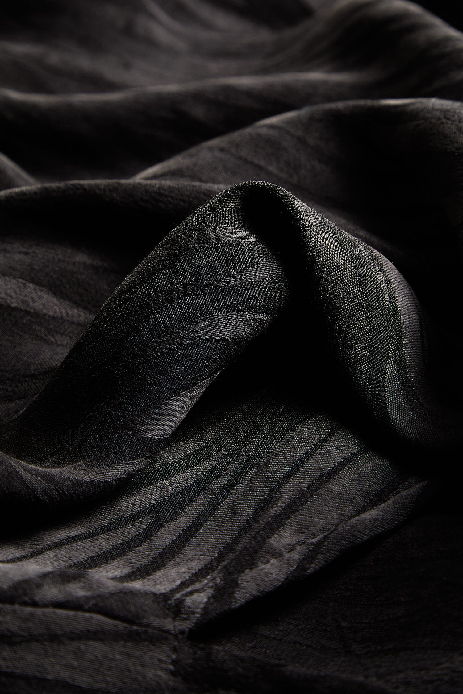 Up-close view of the black and dark gray fabric highlights its wavy, textured pattern. The subtle light reflections and folds offer a contemporary appearance. Crafted from silky-feel fabric, the Abstract Lines Jacquard Bubble Trouser exudes elegance in its design.