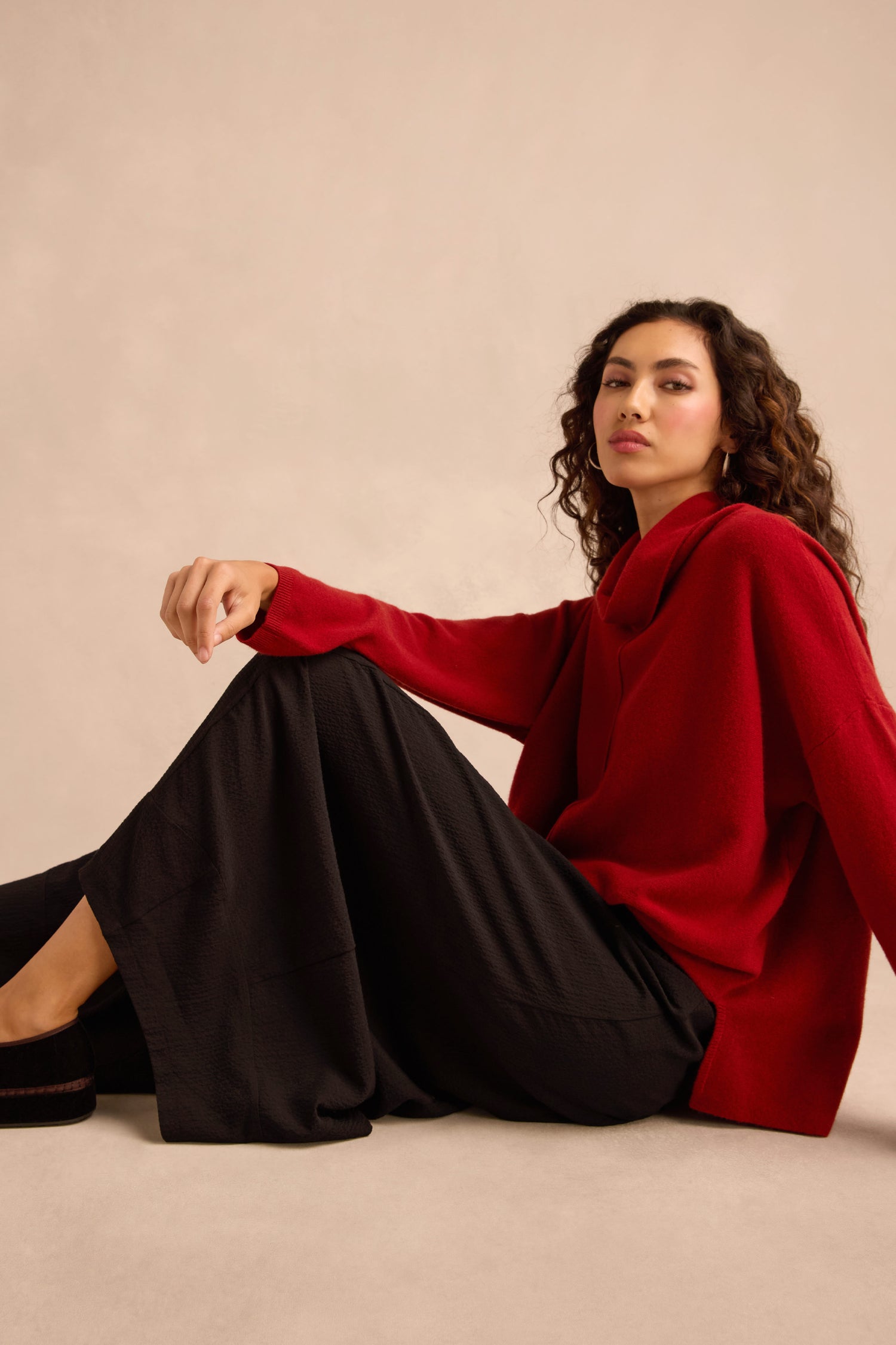 A person with long, curly hair sits on the floor wearing a red sweater and Fine Crinkle Barrel Trousers with a cropped length against a neutral background.