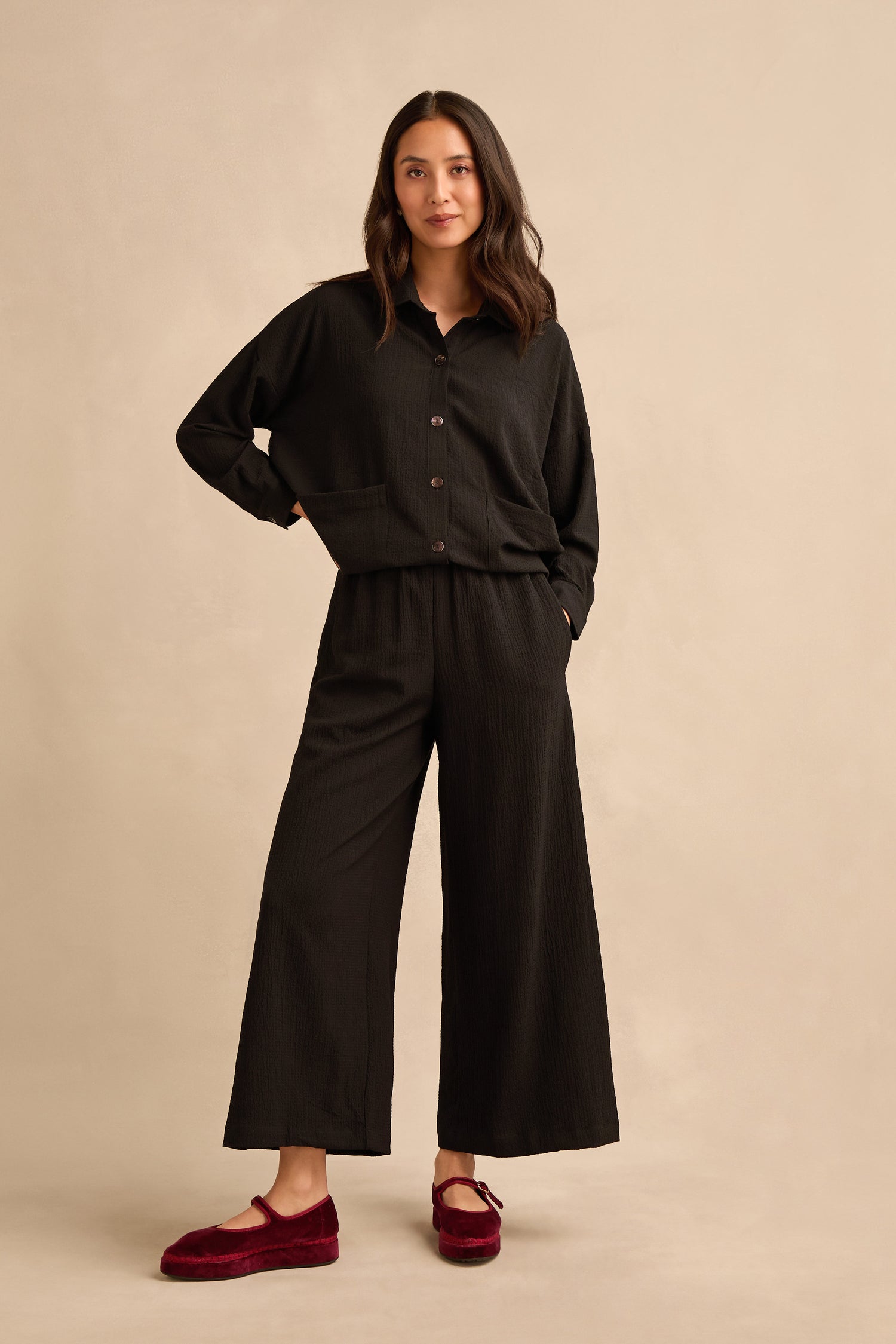 A woman stands against a beige background wearing a loose black button-up top, Mini Crinkle Viscose Wide Leg Trousers with an elasticated waist, and red slippers. One hand rests in her pocket; her hair is loose and wavy.