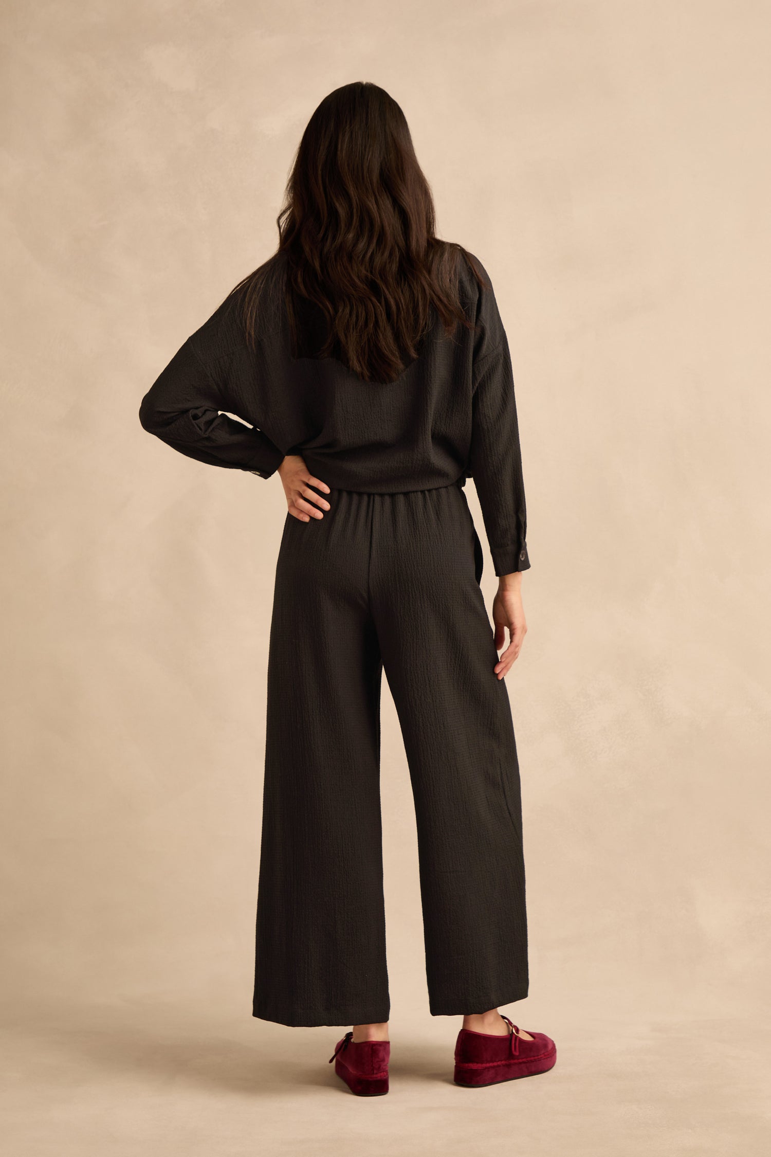 A person with long, wavy brown hair stands facing away against a beige background, wearing the Mini Crinkle Viscose Wide Leg Trouser in black, paired with a loose black top and red shoes.