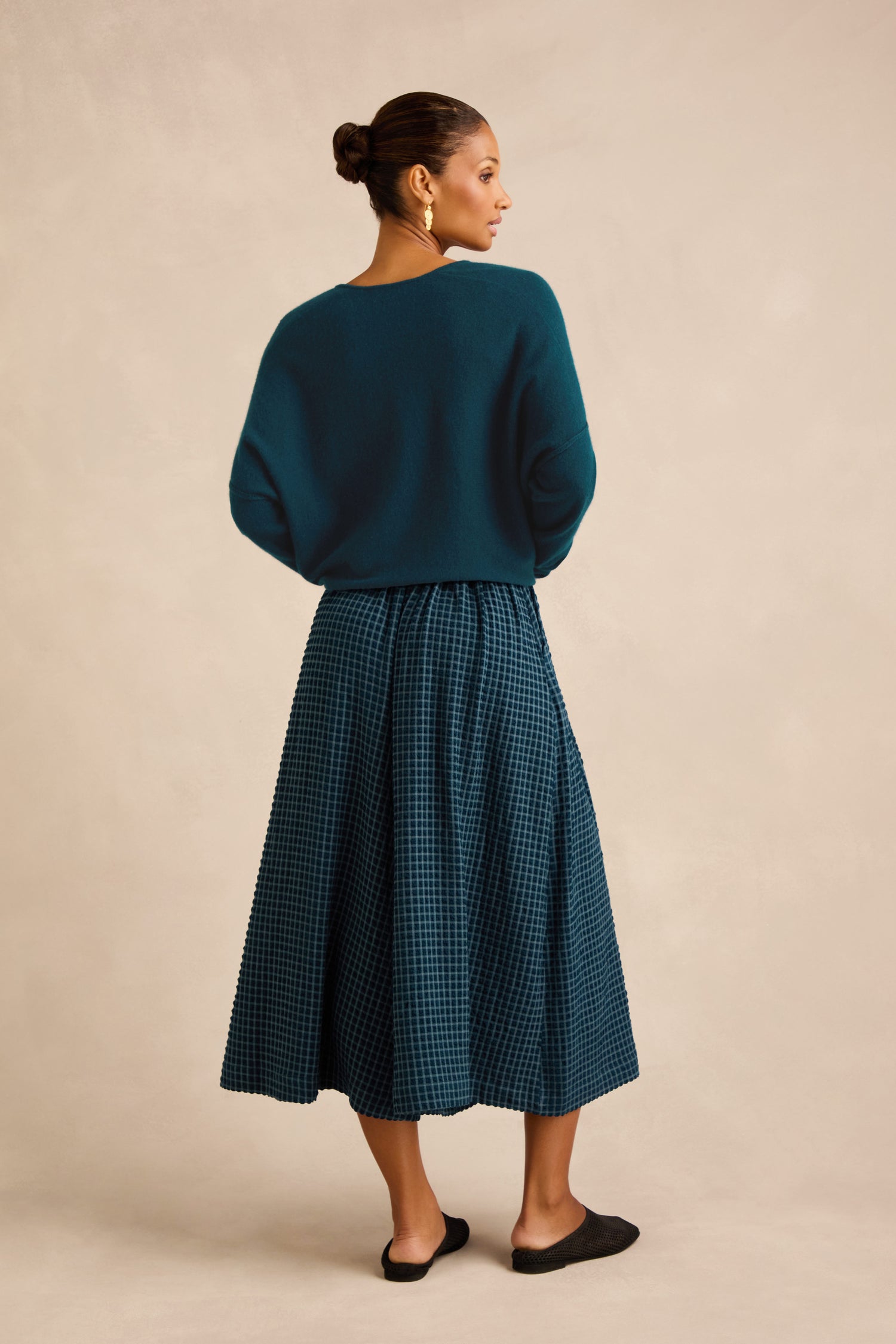 Woman standing with back turned, wearing a teal sweater, Seersucker Squares Wide Trousers styled as a midi skirt, black flats, and gold earrings, with her hair in a neat bun against a neutral background.