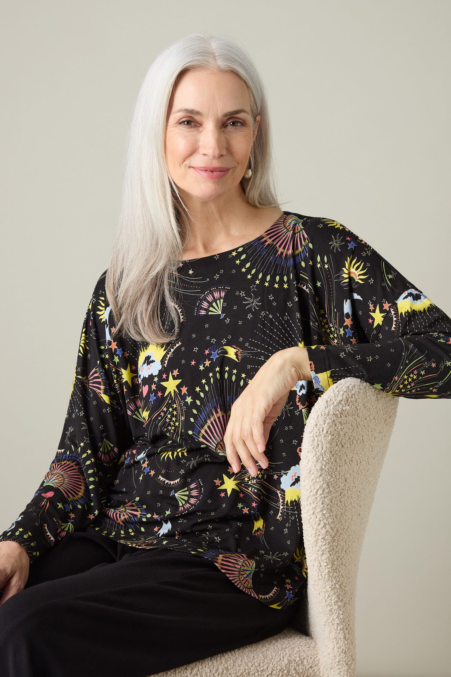A woman with long gray hair sits in a chair, wearing the Celestial Dreams Jersey Boxy Top adorned with colorful celestial patterns.