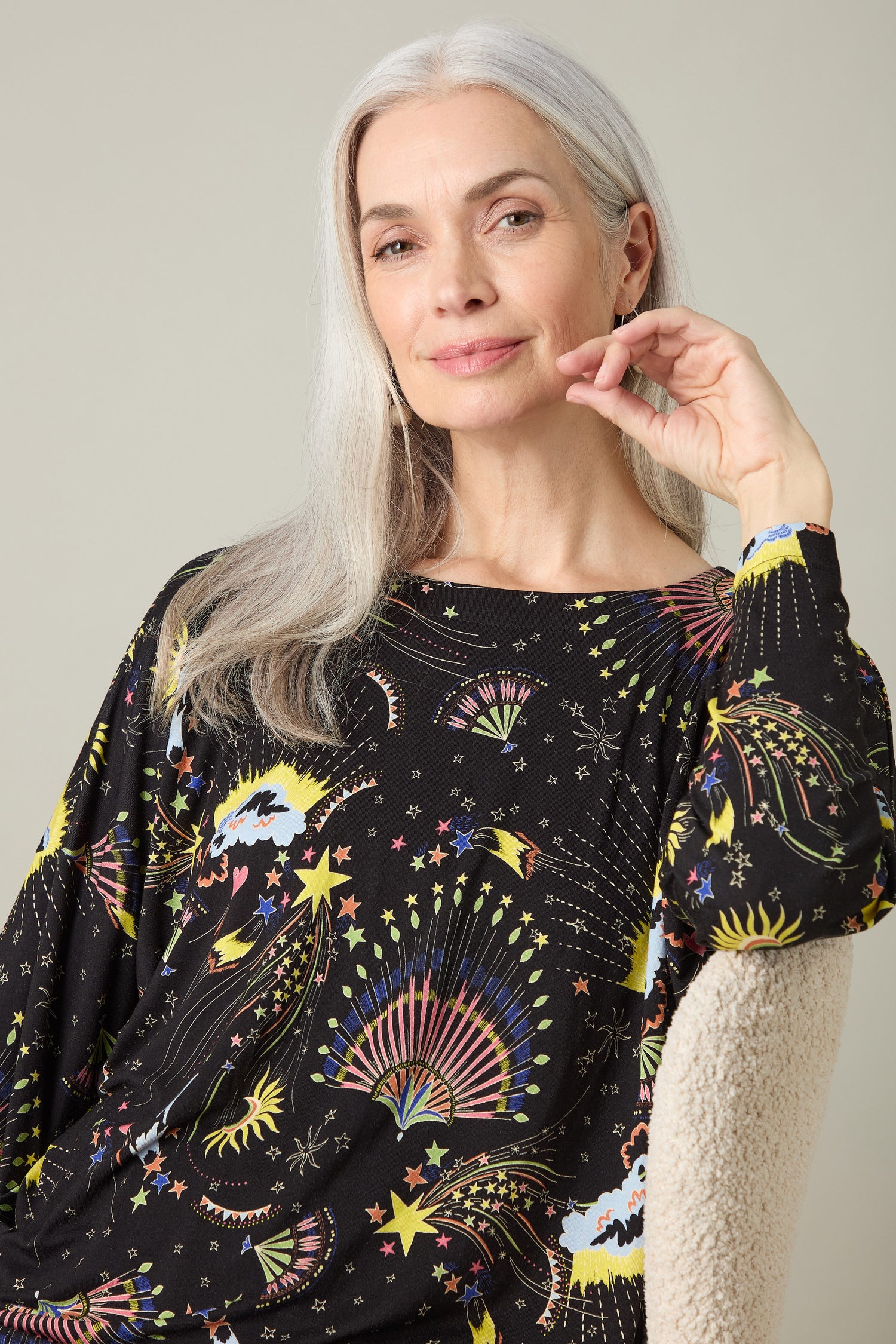 An older adult with long gray hair sits with a slight smile, resting her chin on her hand against a neutral background while wearing the vibrant Celestial Dreams Jersey Boxy Top.