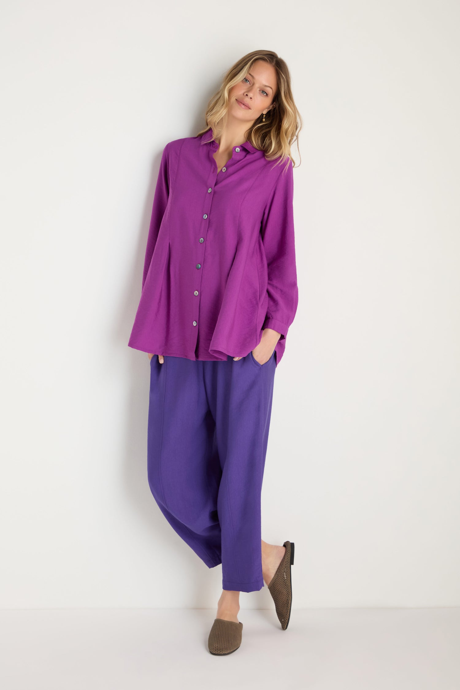 A woman stands against a white wall wearing the Textured Viscose Pleat Shirt in purple with matching purple pants and brown slip-on shoes, one hand in her pocket.