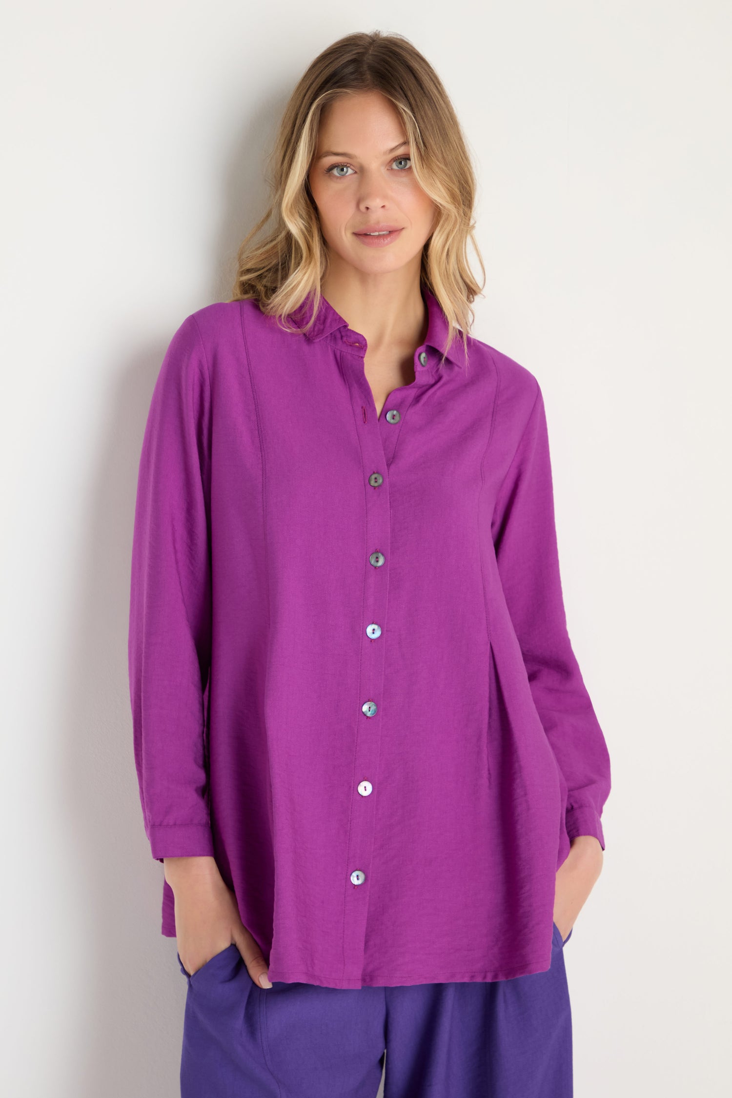 A woman stands against a white wall wearing the Textured Viscose Pleat Shirt and matching purple pants, with her hands in her pockets.