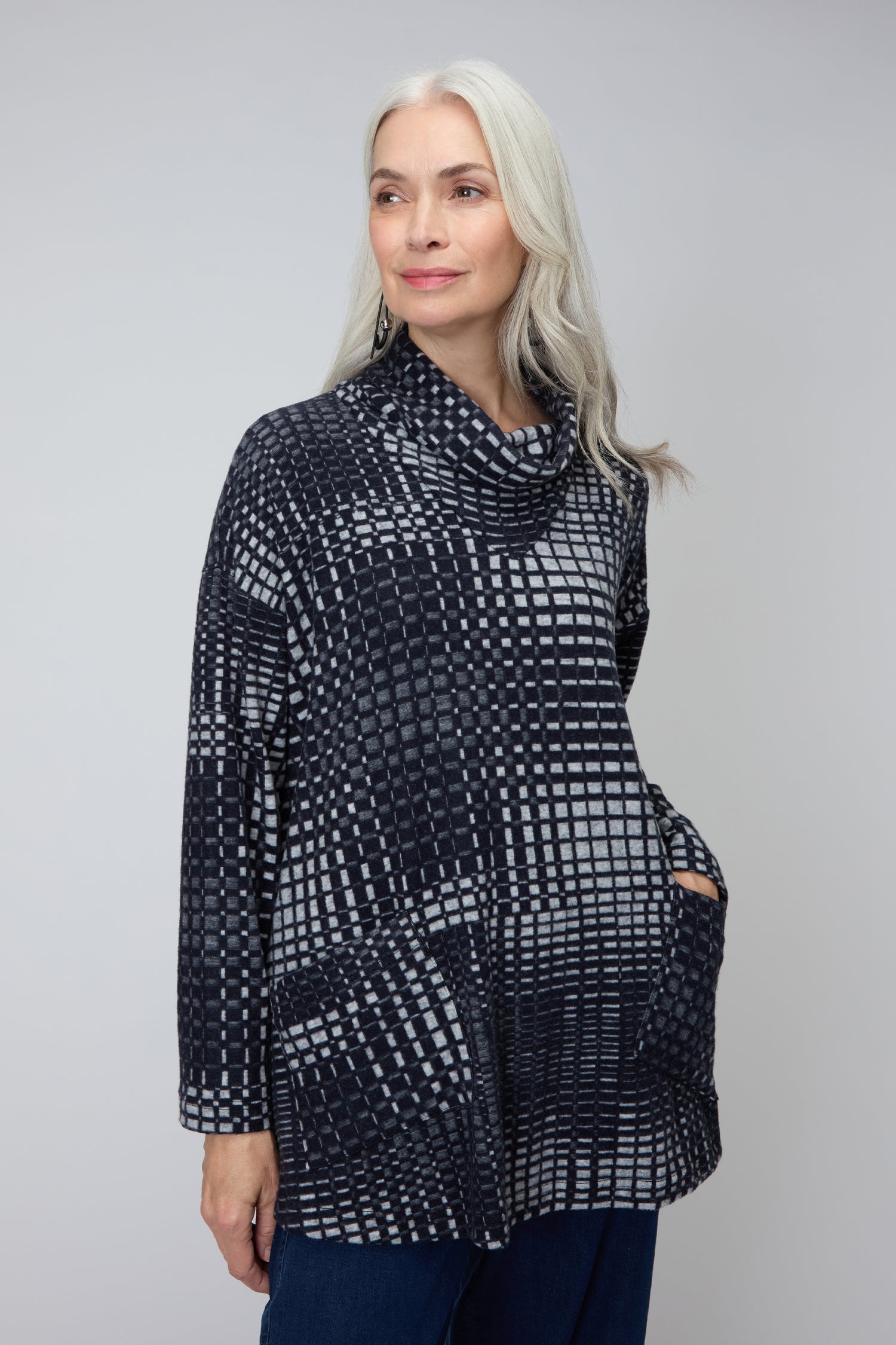 Woman with long gray hair wearing the Illusion Check Jersey Tunic with a cowl collar and front pockets, standing against a plain light gray background.