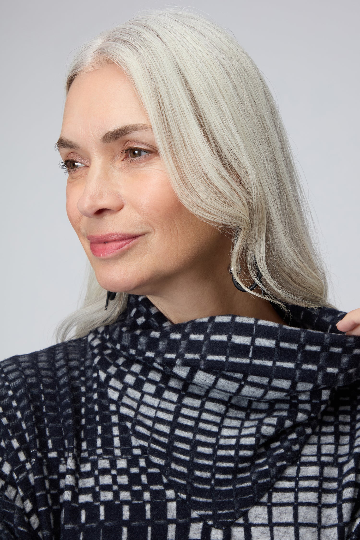 A woman with long gray hair smiles softly to the side, holding the cowl collar of her Illusion Check Jersey Tunic in geometric blue and gray jacquard.