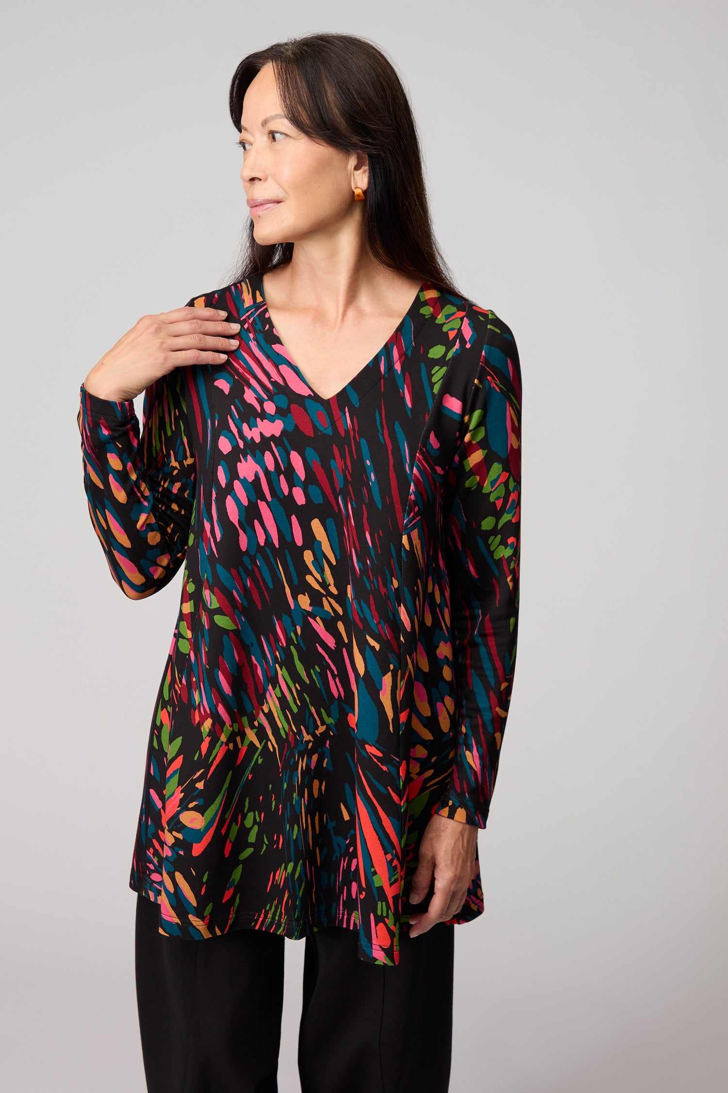 A woman stands against a plain background, wearing the Skyfire Jersey Top in a multicolored abstract print and black pants, looking to the side.
