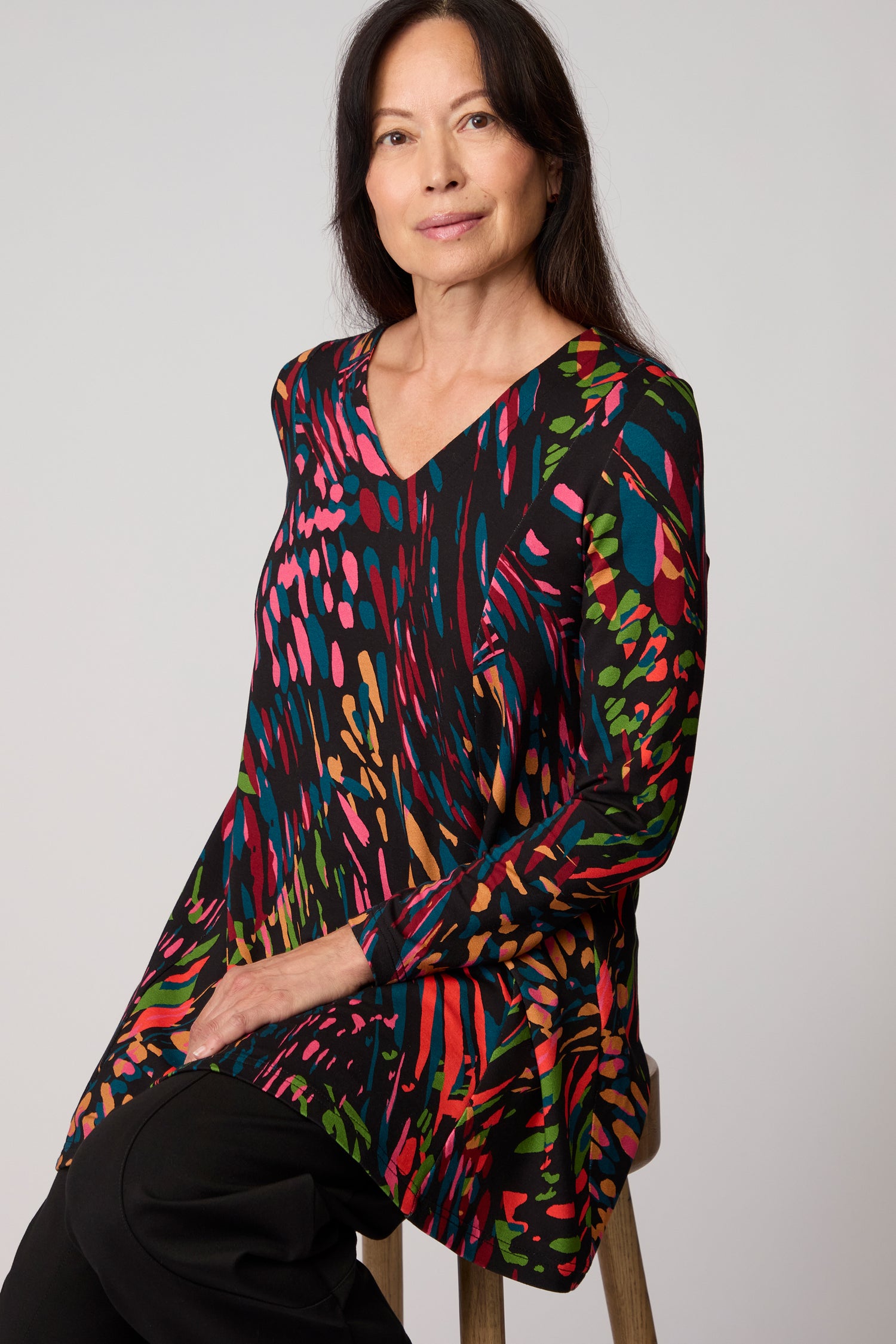 A woman with long dark hair sits on a stool, wearing the Skyfire Jersey Top—a black jersey with a colorful abstract print—and black pants, set against a plain light background.