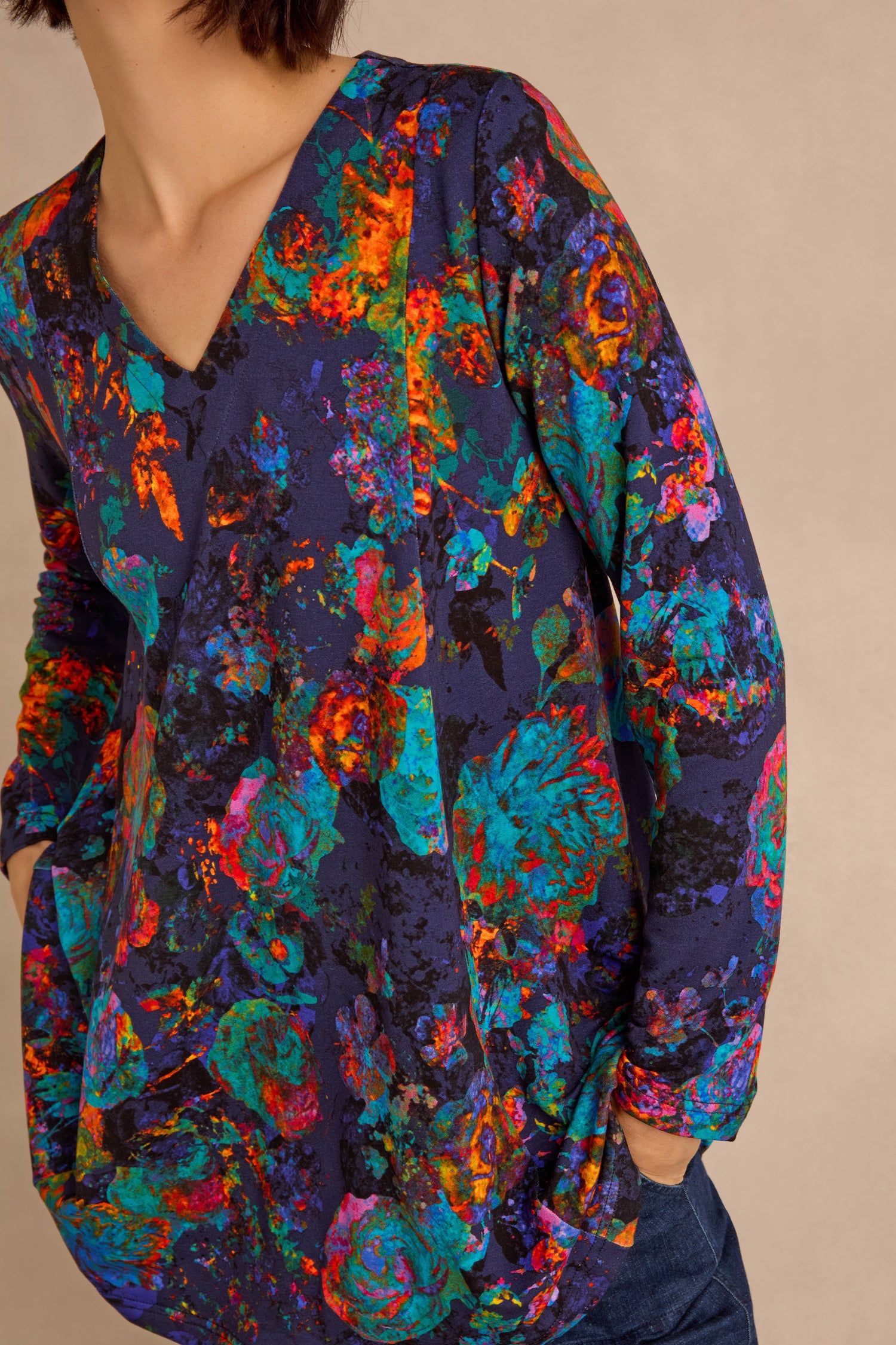 Wearing the Midnight Floral Jersey Tunic, a long-sleeved, V-neck top with a colorful abstract floral pattern, paired with blue jeans and standing against a neutral background.