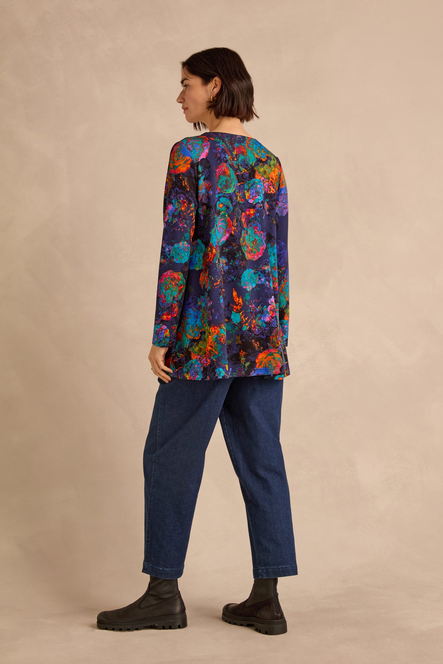 A person stands sideways against a beige background, wearing the Midnight Floral Jersey Tunic with long sleeves, a colorful floral pattern, and a flattering fit-and-flare silhouette, paired with blue jeans and black boots.