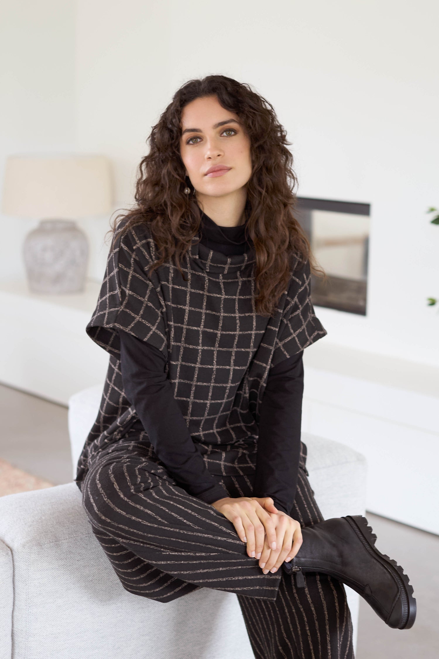 A woman with curly hair sits on a light-colored sofa, wearing a Broken Check Jersey Tabard with black boots, in a minimalist room.