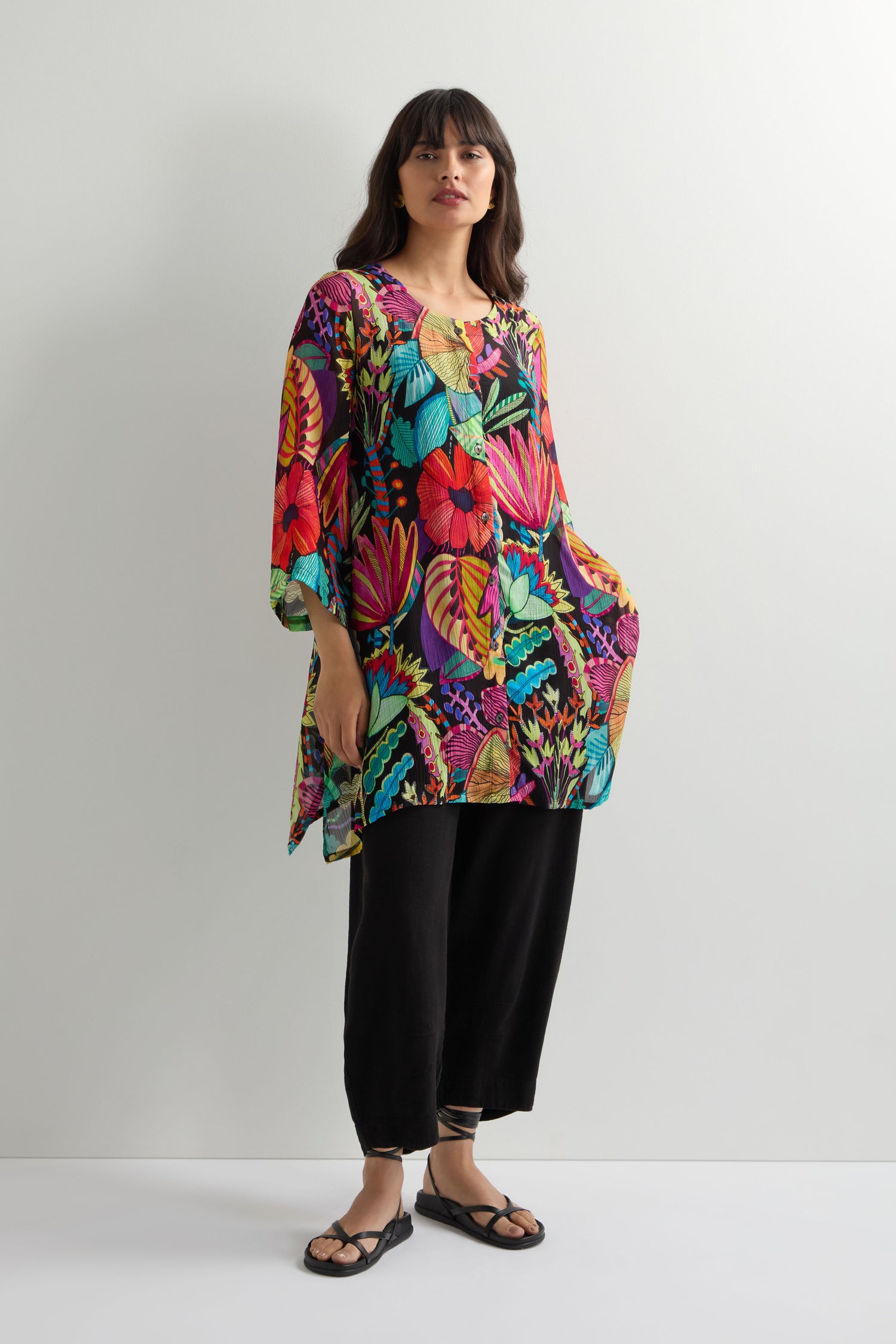 A woman stands against a plain wall, wearing the Fantasy Forest Georgette Shirt featuring a rainforest print, paired with loose black pants and black sandals.